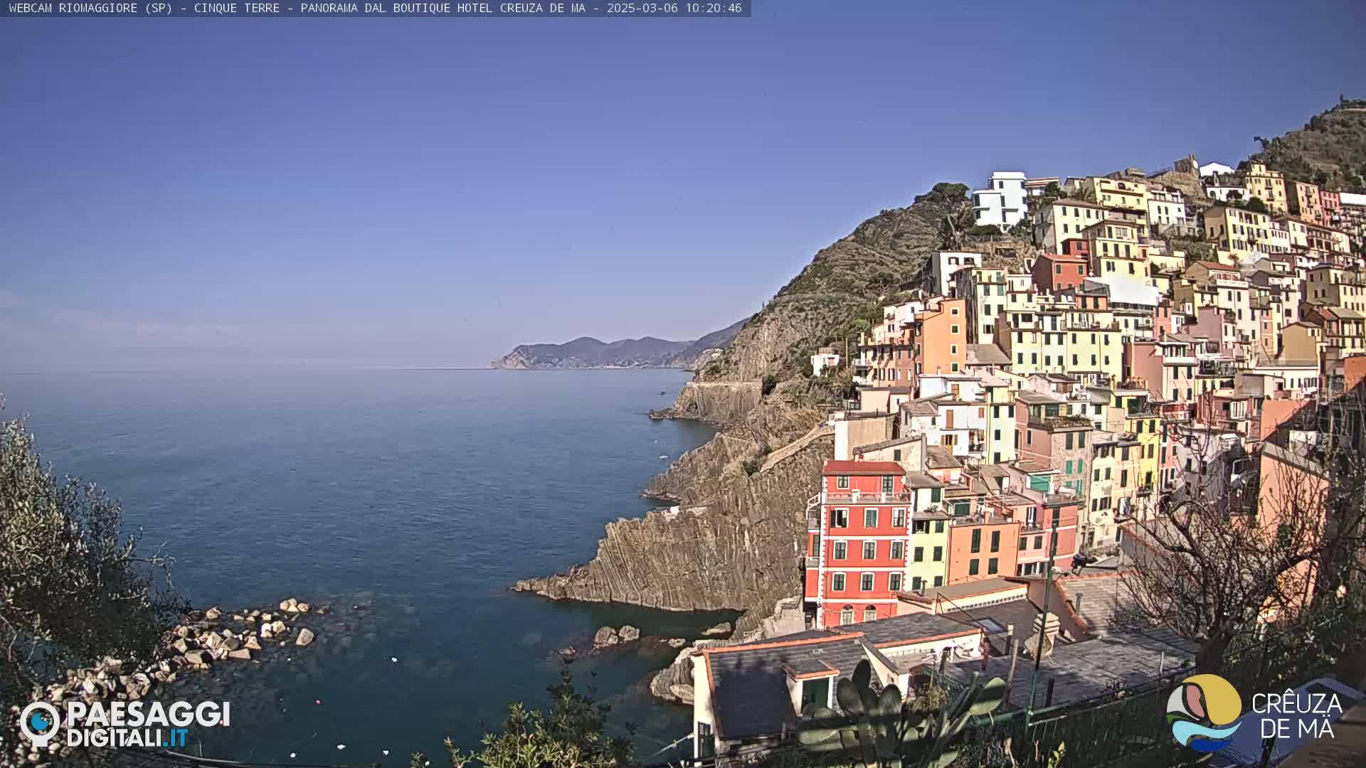 A colorful hillside village clings to a rocky coastline overlooking a calm blue sea under a clear, sunny sky.