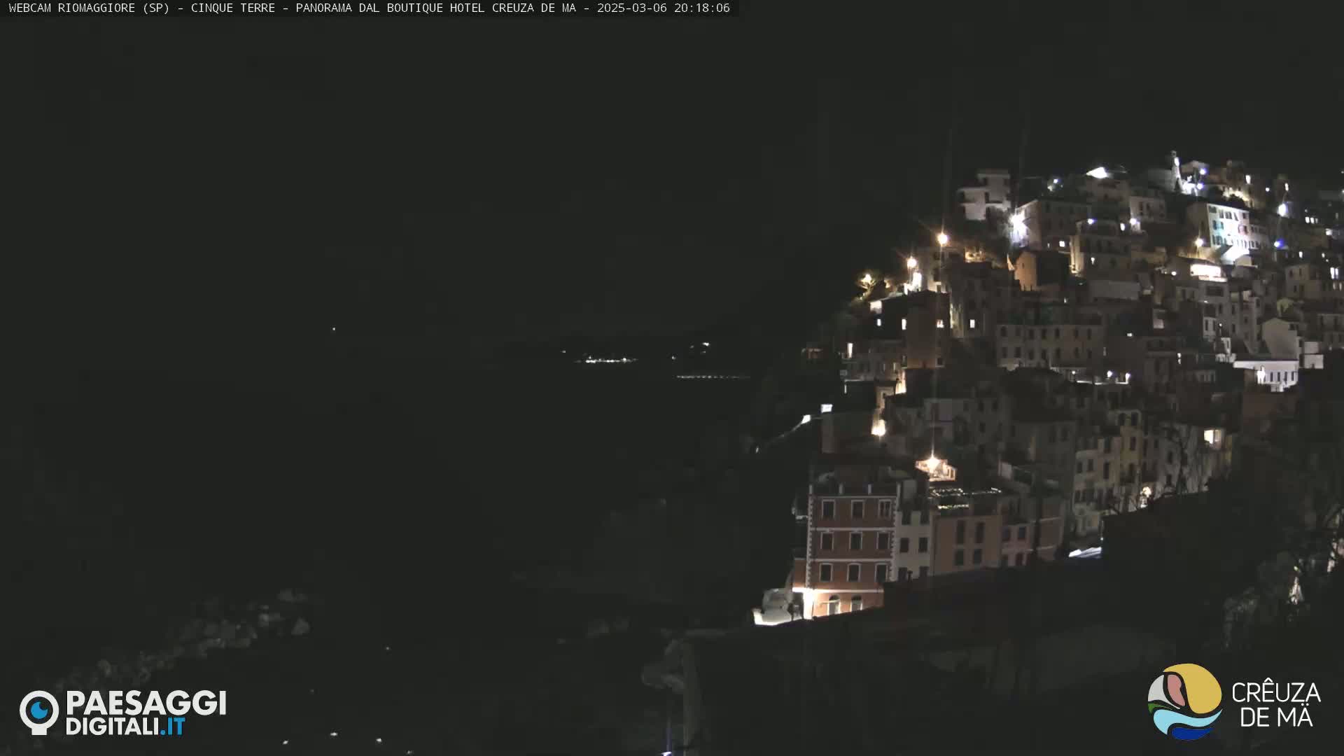 A nighttime view of a coastal town with many buildings lit up, overlooking a dark body of water under a clear sky.