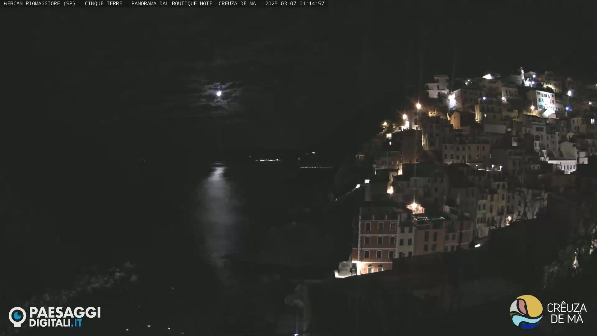 A nighttime view of a coastal town illuminated by lights, with a bright moon visible in a partly cloudy sky over calm water.