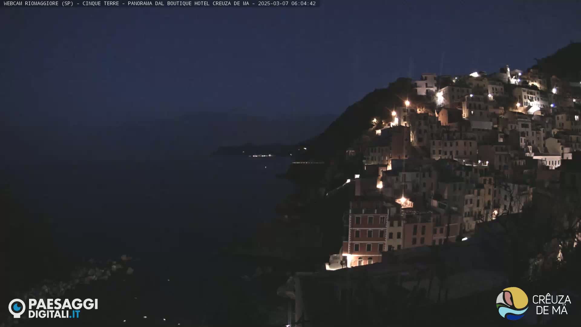 A hillside village is illuminated at night, overlooking a dark body of water under a clear, dark sky.