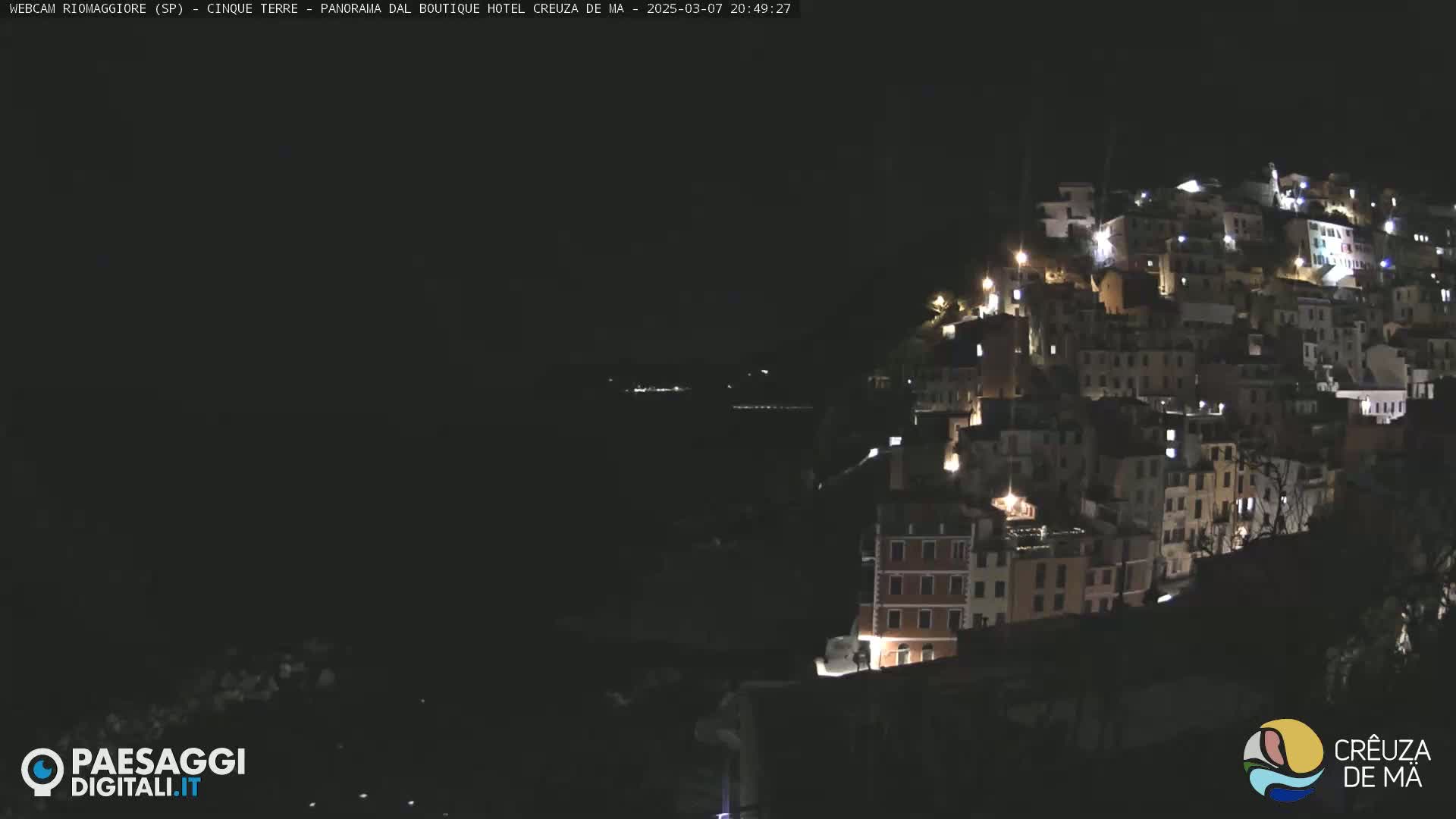 A nighttime view of a hillside town with many illuminated buildings, overlooking a dark body of water under a clear, dark sky.