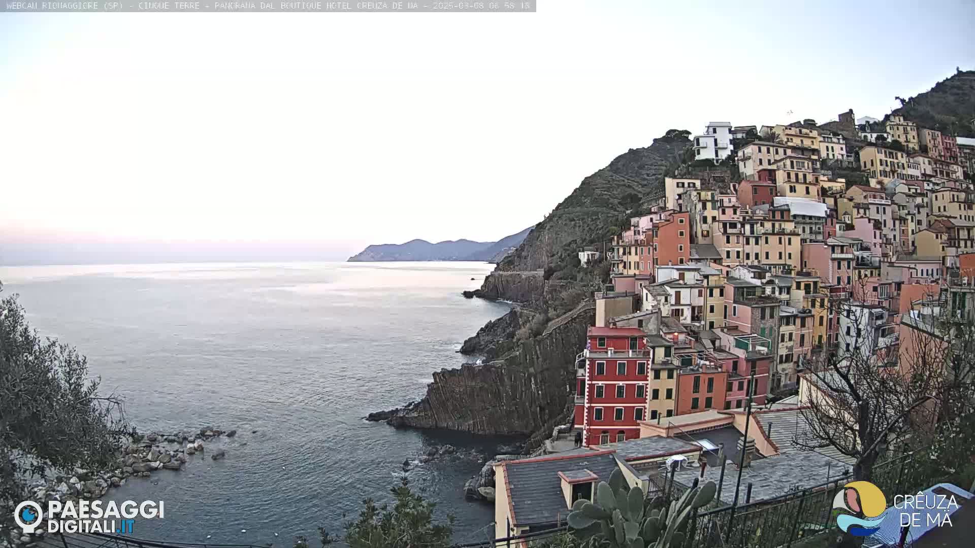 A coastal town with colorful buildings clinging to a steep hillside overlooks a calm sea under a pale sky.