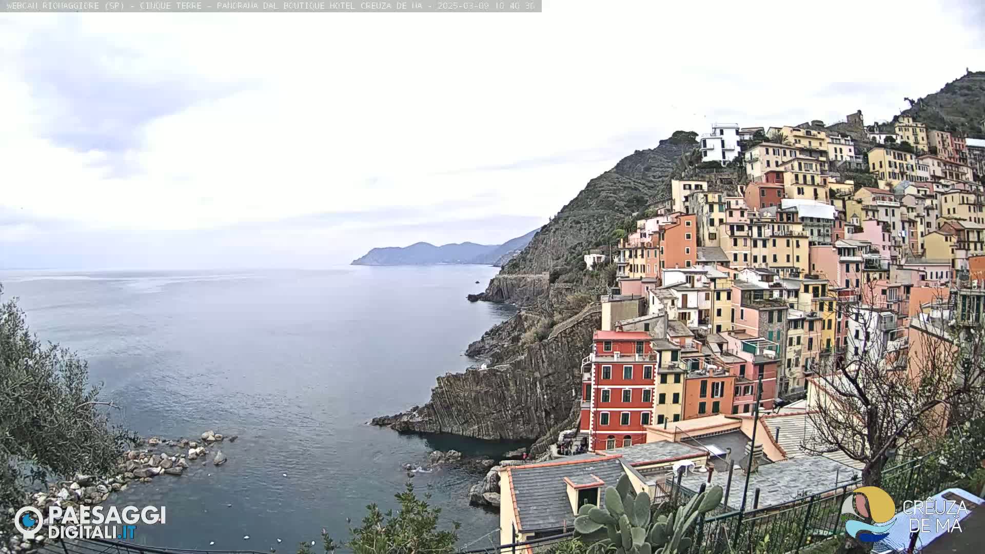 A coastal village of colorful buildings clinging to a steep hillside overlooks a calm sea under a cloudy sky.