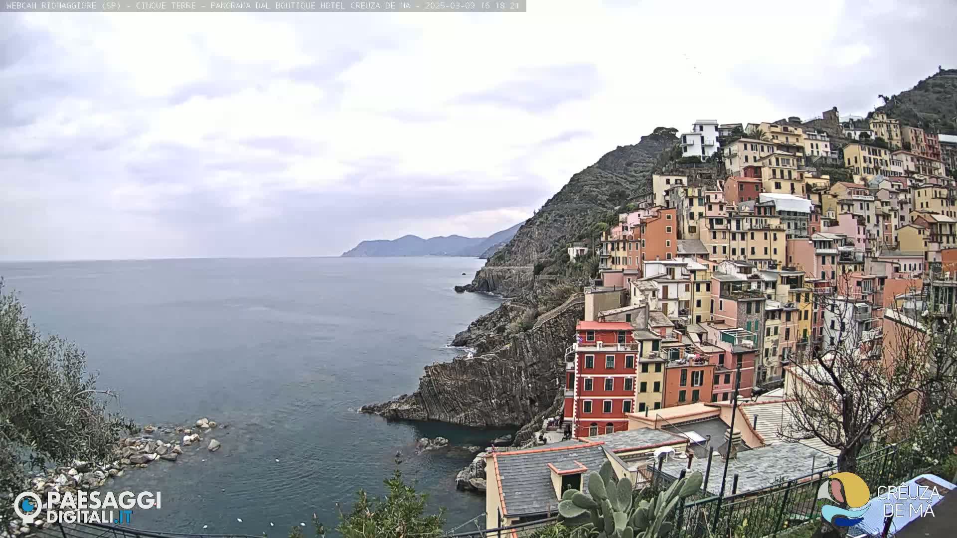 A coastal village of colorful buildings climbs a steep hillside overlooking a calm sea under an overcast sky.