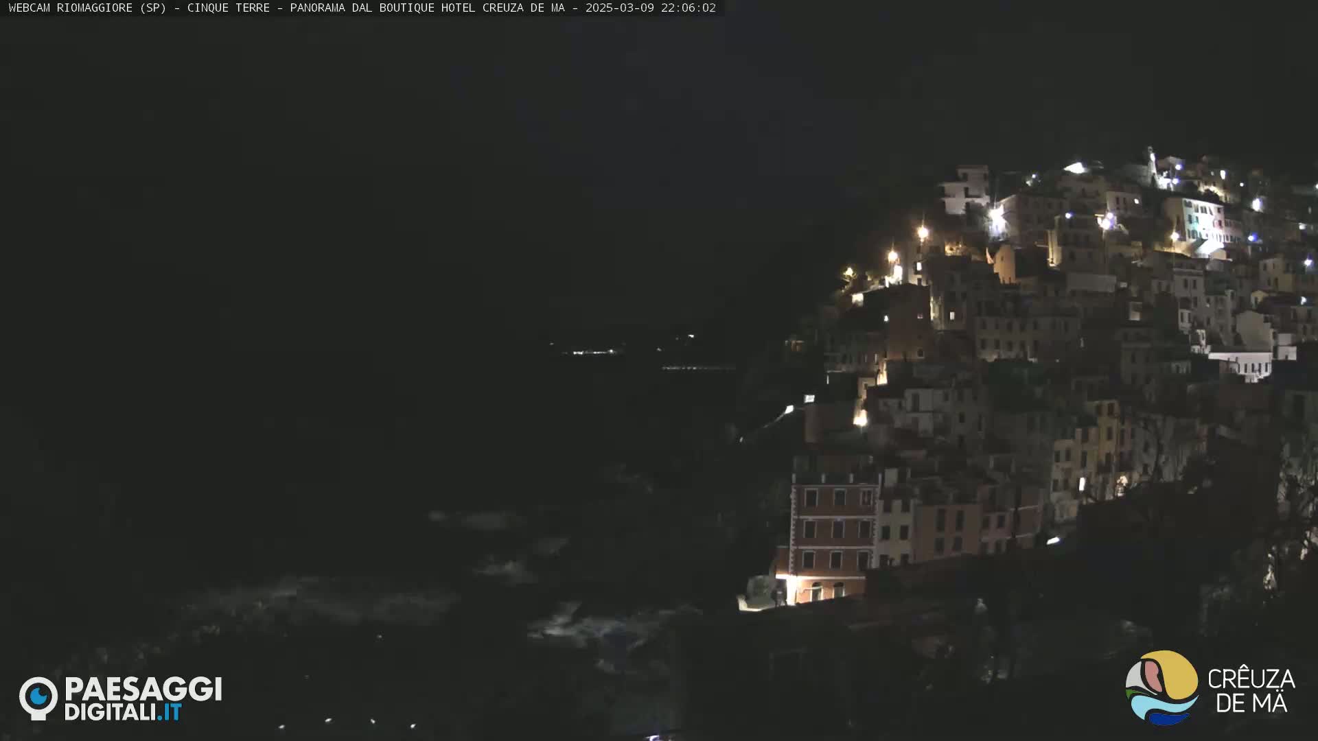 A nighttime view of a hilltop village illuminated by lights, overlooking a dark body of water.
