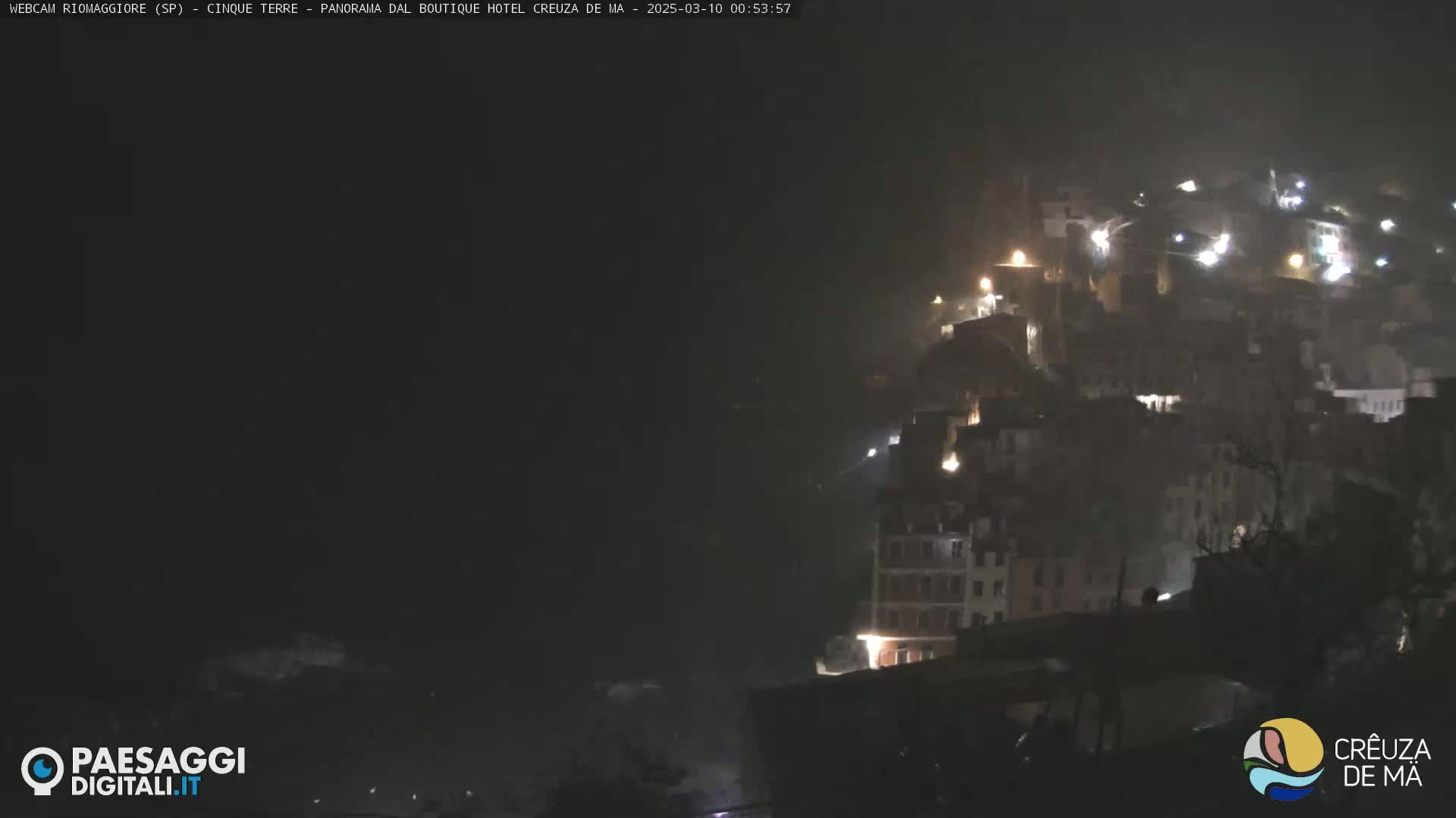 A nighttime view of a town on a hillside shrouded in heavy rain or fog, with the lights of buildings visible through the precipitation.