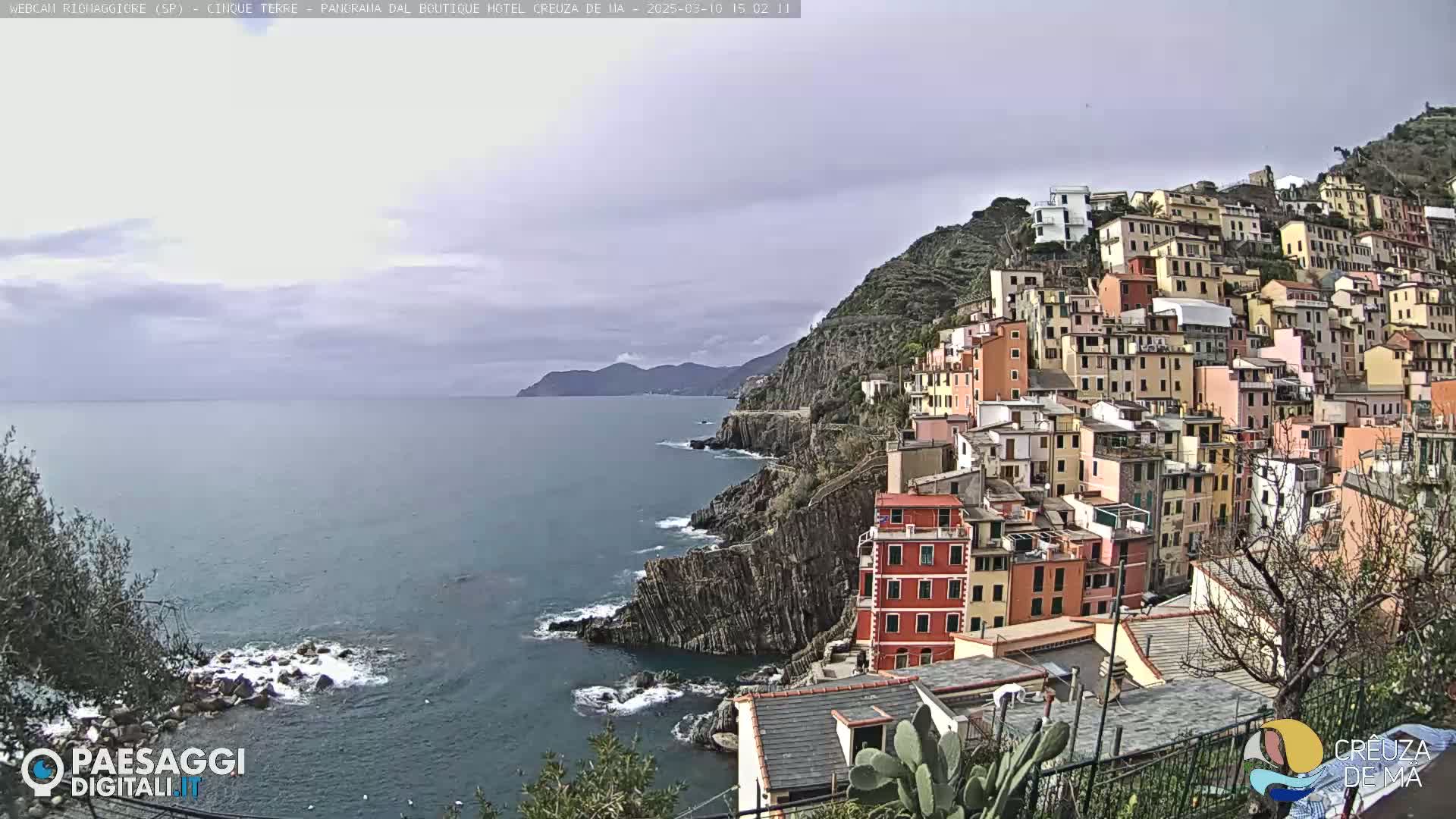 A coastal town with colorful buildings stacked on a steep hillside overlooks a calm sea under a cloudy sky.