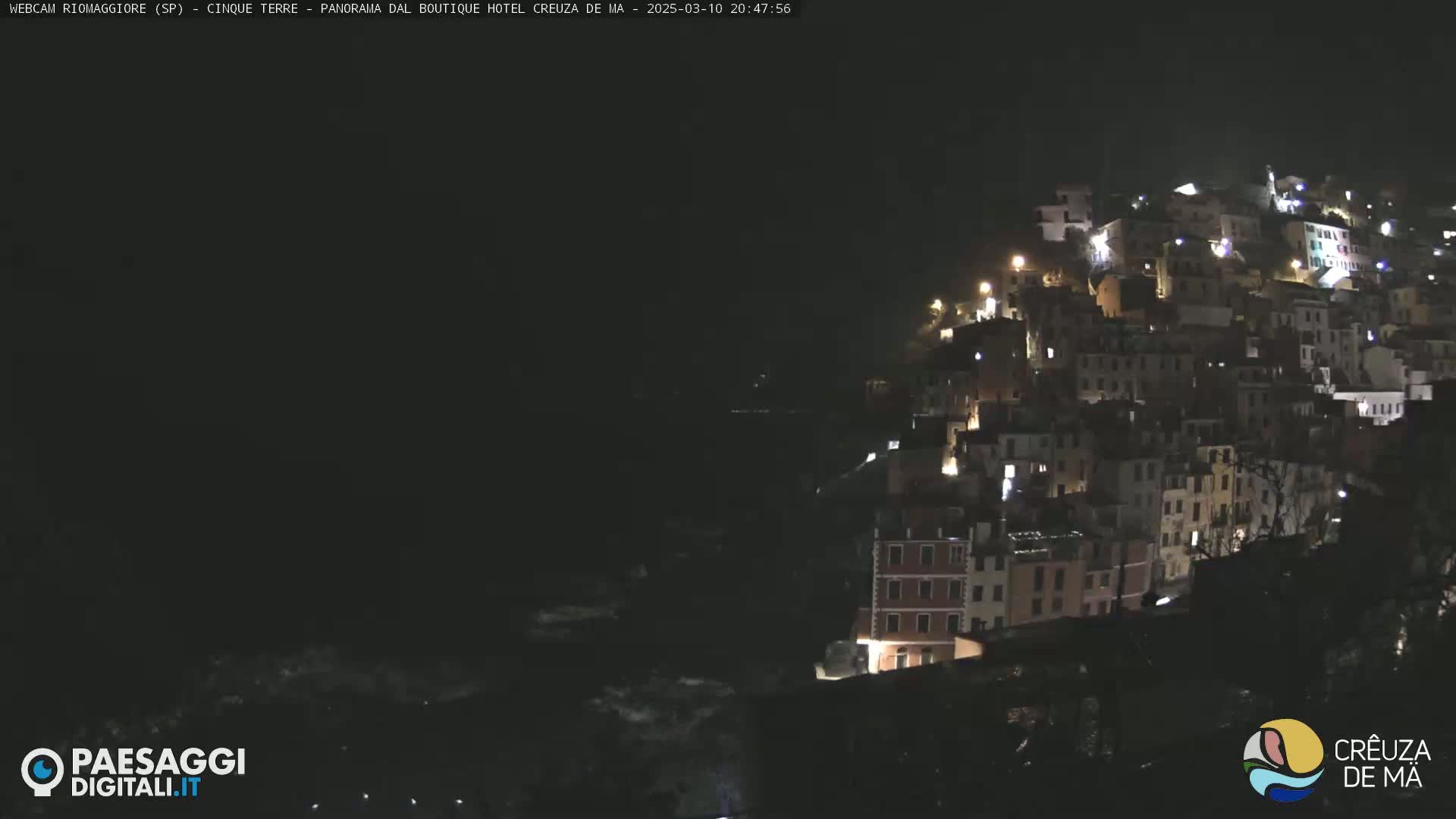 A nighttime view of a hillside town with many lit windows, overlooking a dark body of water.