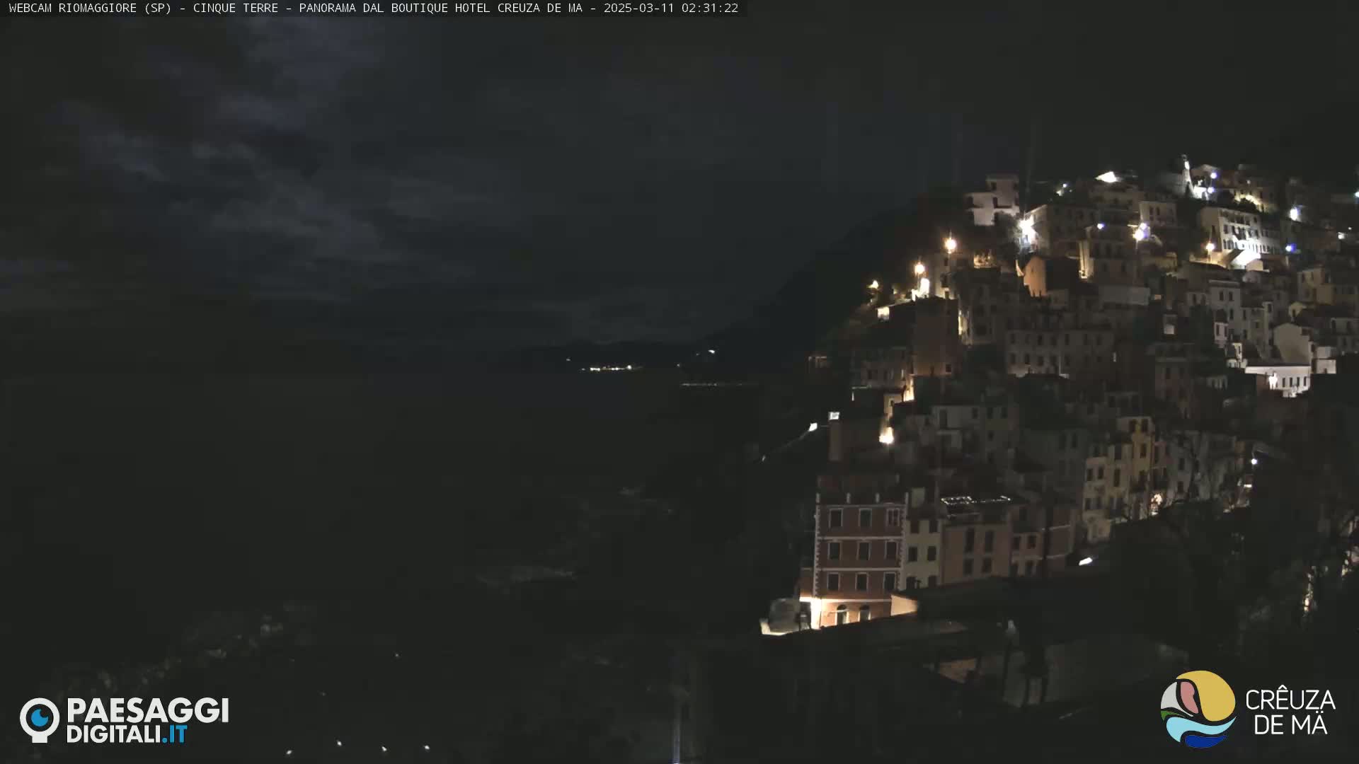 A nighttime, slightly cloudy view of a hilltop village illuminated by lights, overlooking a dark body of water.