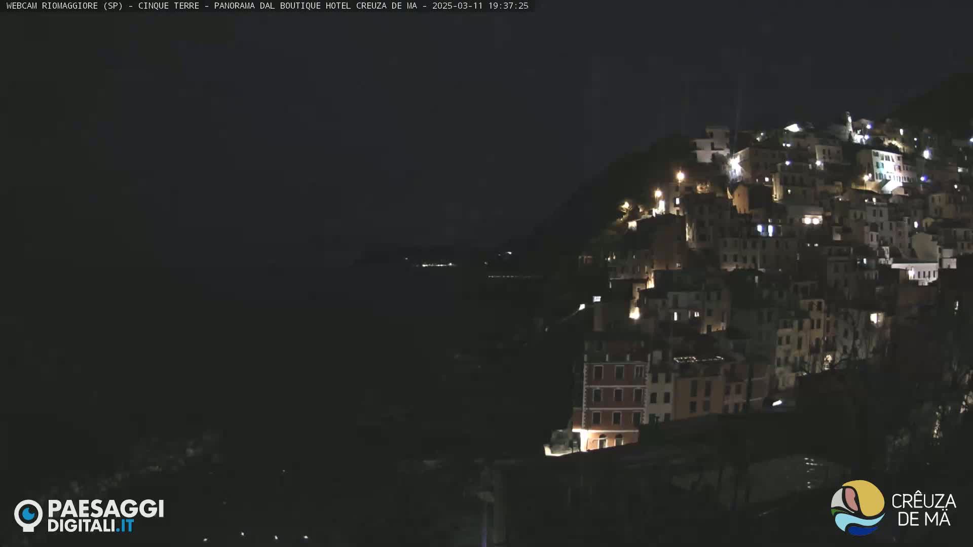 A nighttime, seaside village clings to a hillside, its buildings illuminated against a dark ocean.