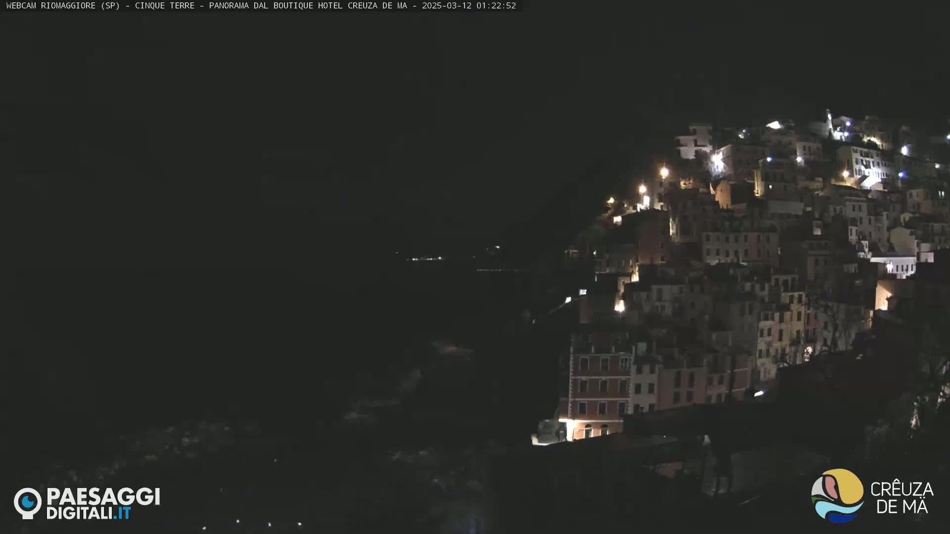 A nighttime view of a hillside town with many lit windows, overlooking a dark body of water.