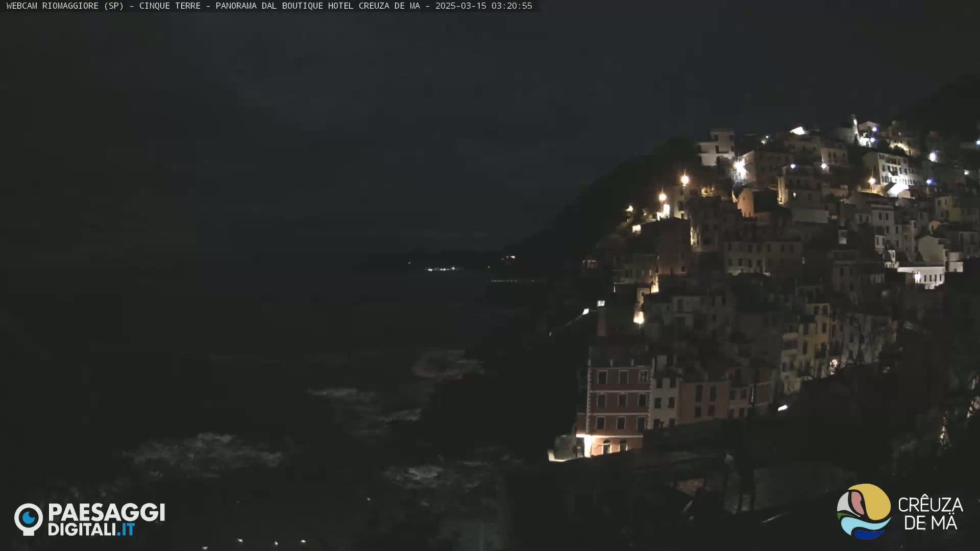 A nighttime view of a coastal town illuminated by lights, with dark, wavy water below.