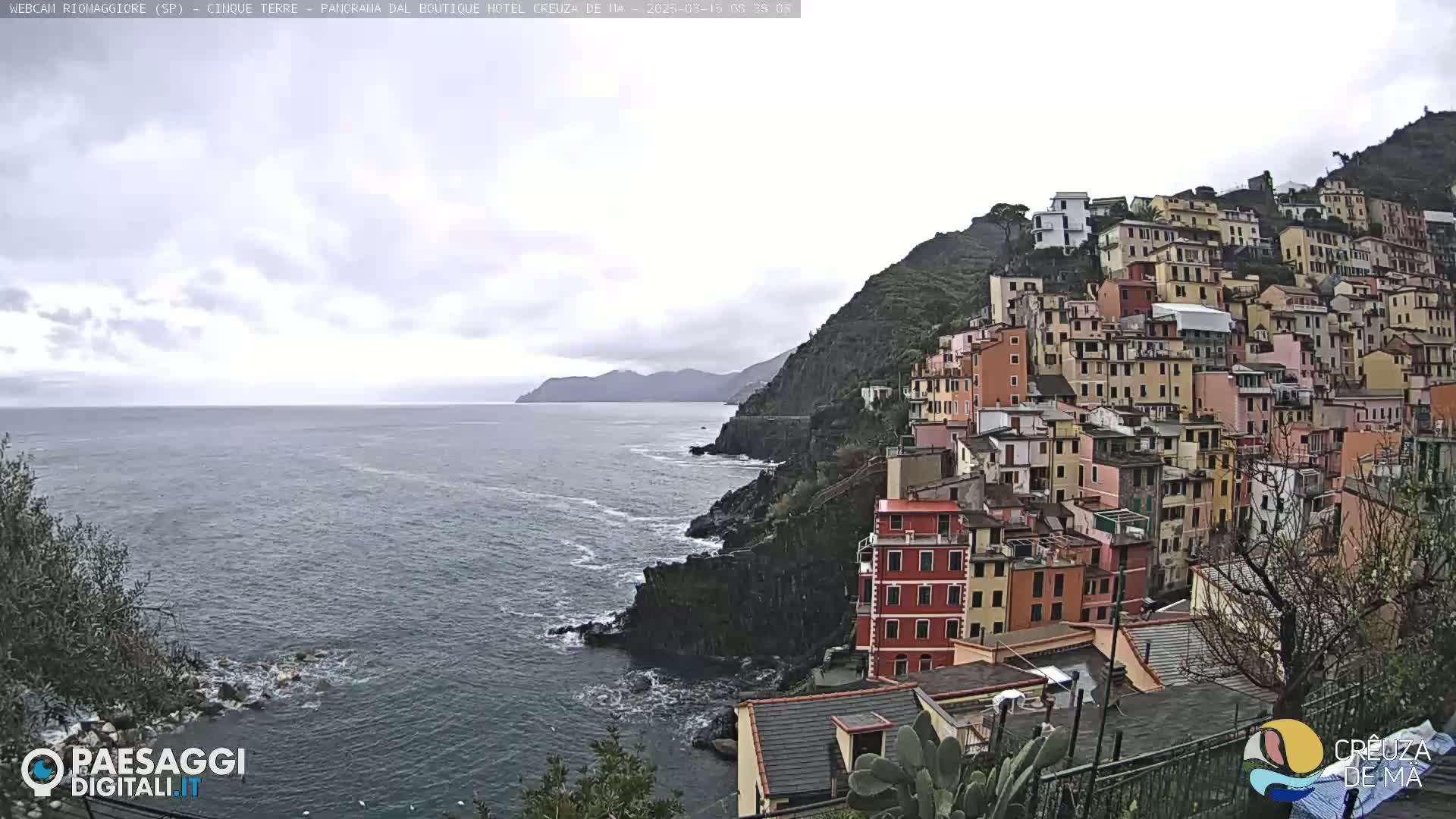 A coastal town with colorful buildings clings to a steep hillside overlooking a calm sea under an overcast sky.