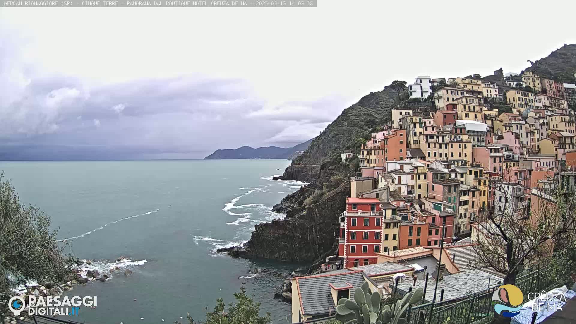 A colorful hillside village clings to a rocky coastline overlooking a calm sea under an overcast sky.