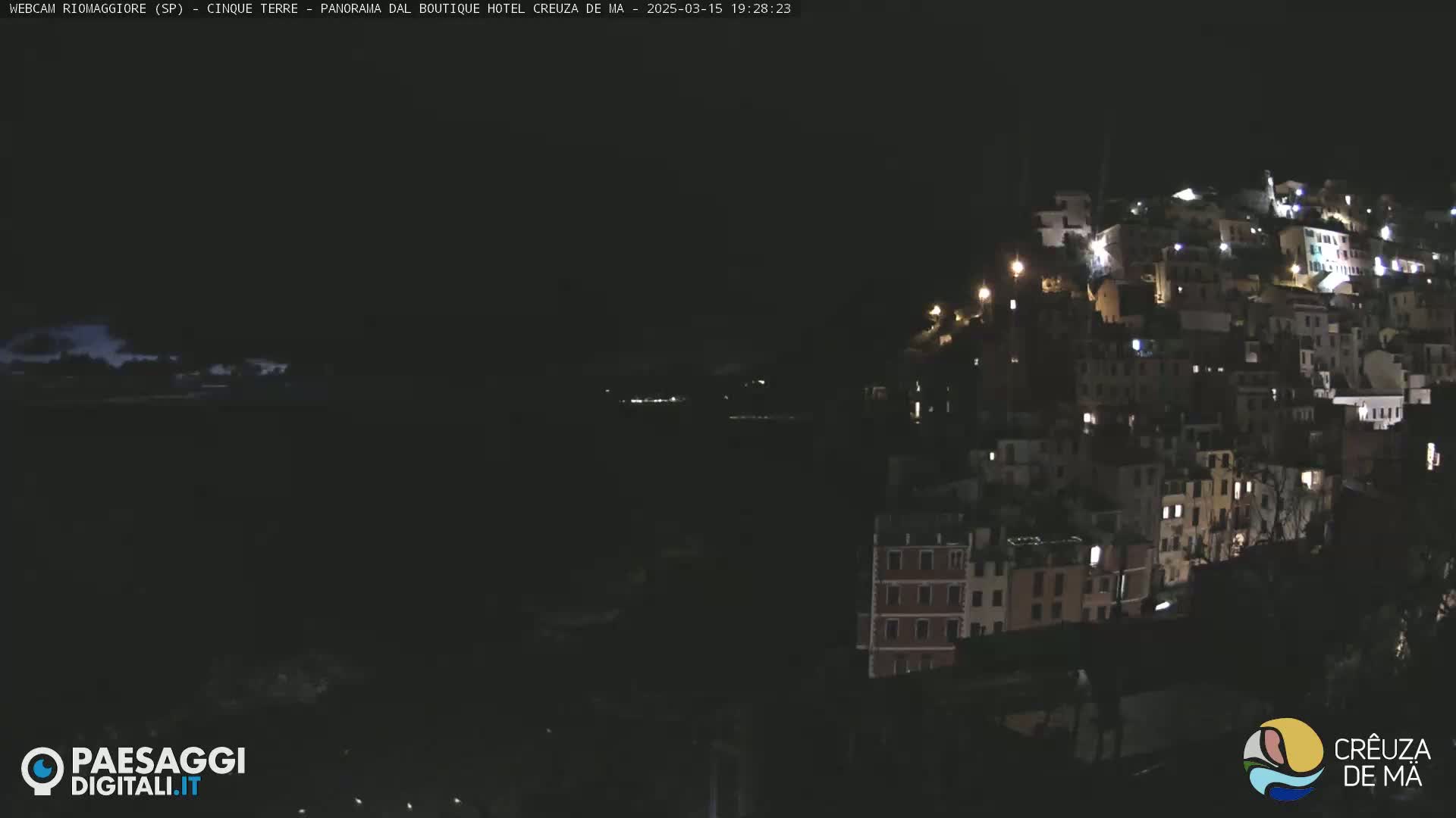 A nighttime view of a hilltop village with numerous lit windows, overlooking a dark body of water under a partly cloudy sky.