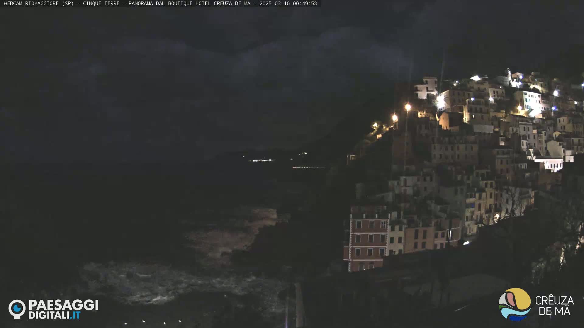 A nighttime view of a hillside town illuminated by lights, overlooking a dark, wavy sea under a cloudy sky.