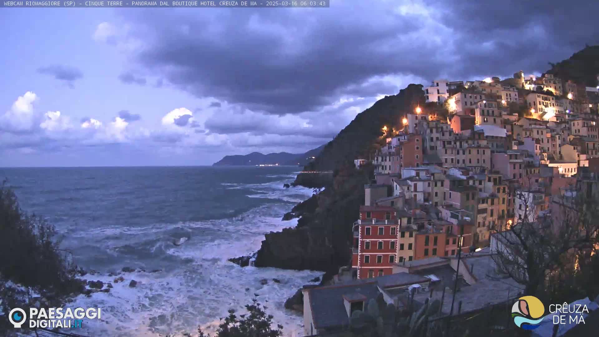 A coastal village with illuminated buildings is perched on a cliff overlooking a turbulent sea under a cloudy twilight sky.