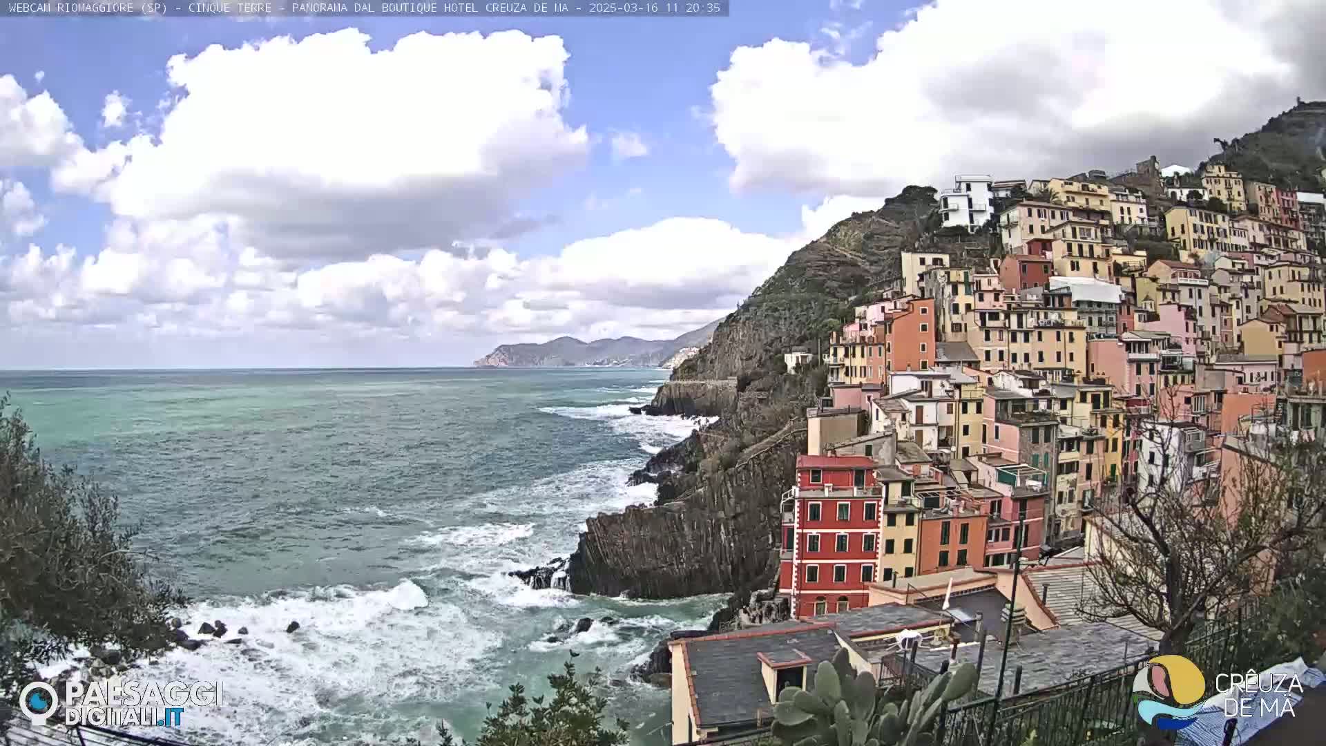 A colorful village clings to a rocky cliffside overlooking a choppy sea under a partly cloudy sky.