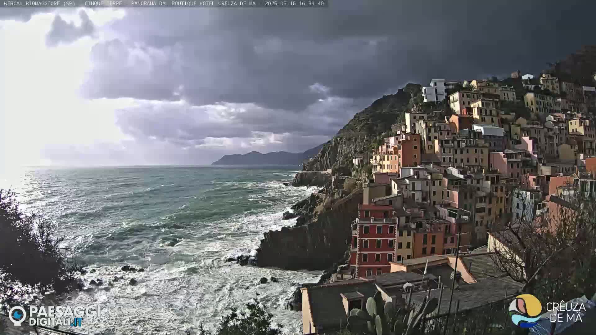A coastal town built on a steep cliffside overlooks a turbulent sea under a partly cloudy sky.