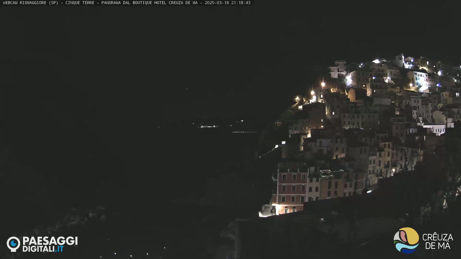 A nighttime view of a hillside town, with many buildings lit up, overlooking a dark body of water.