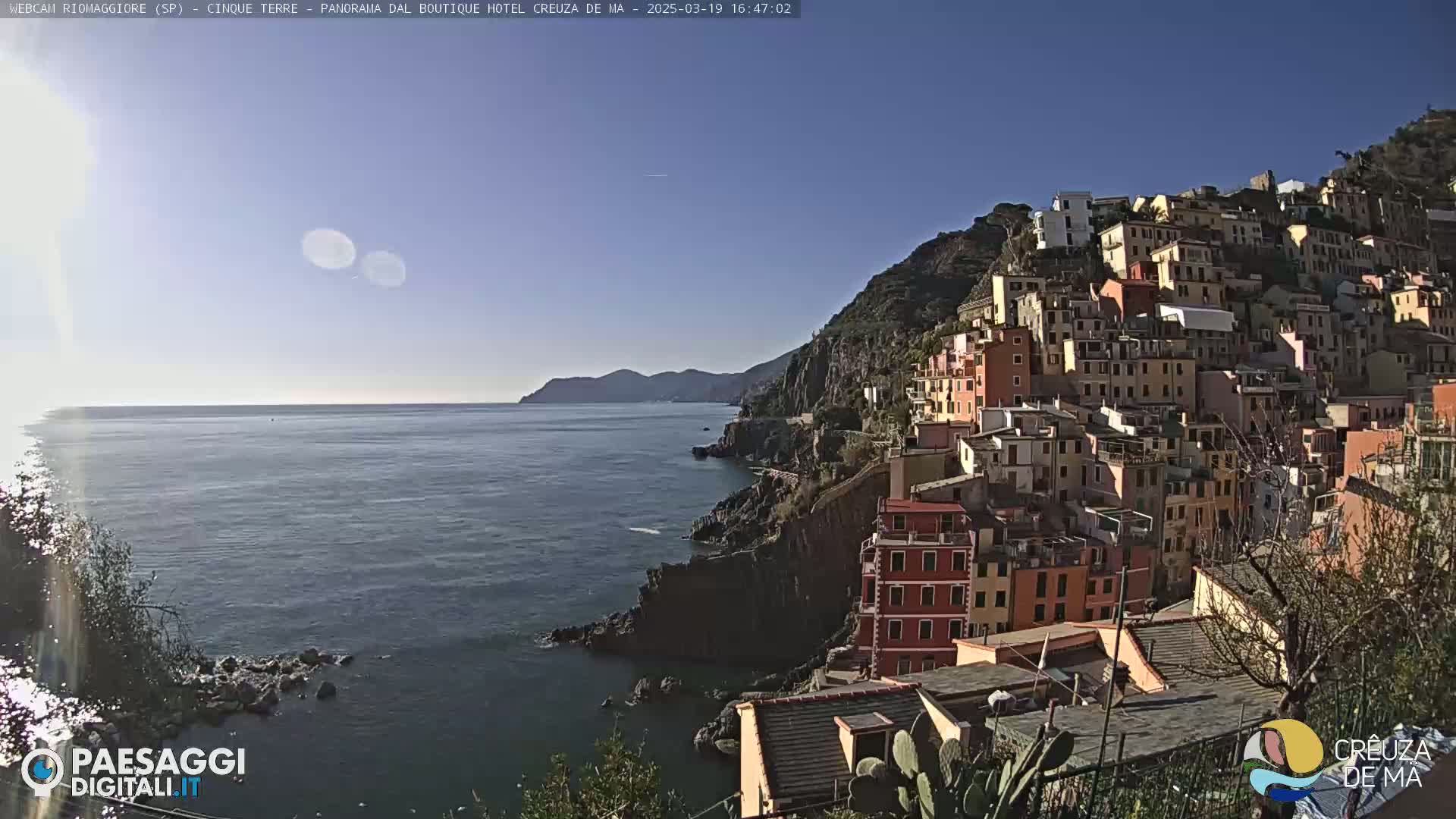 A picturesque coastal town clings to a steep hillside overlooking a calm, blue sea under a clear, sunny sky.