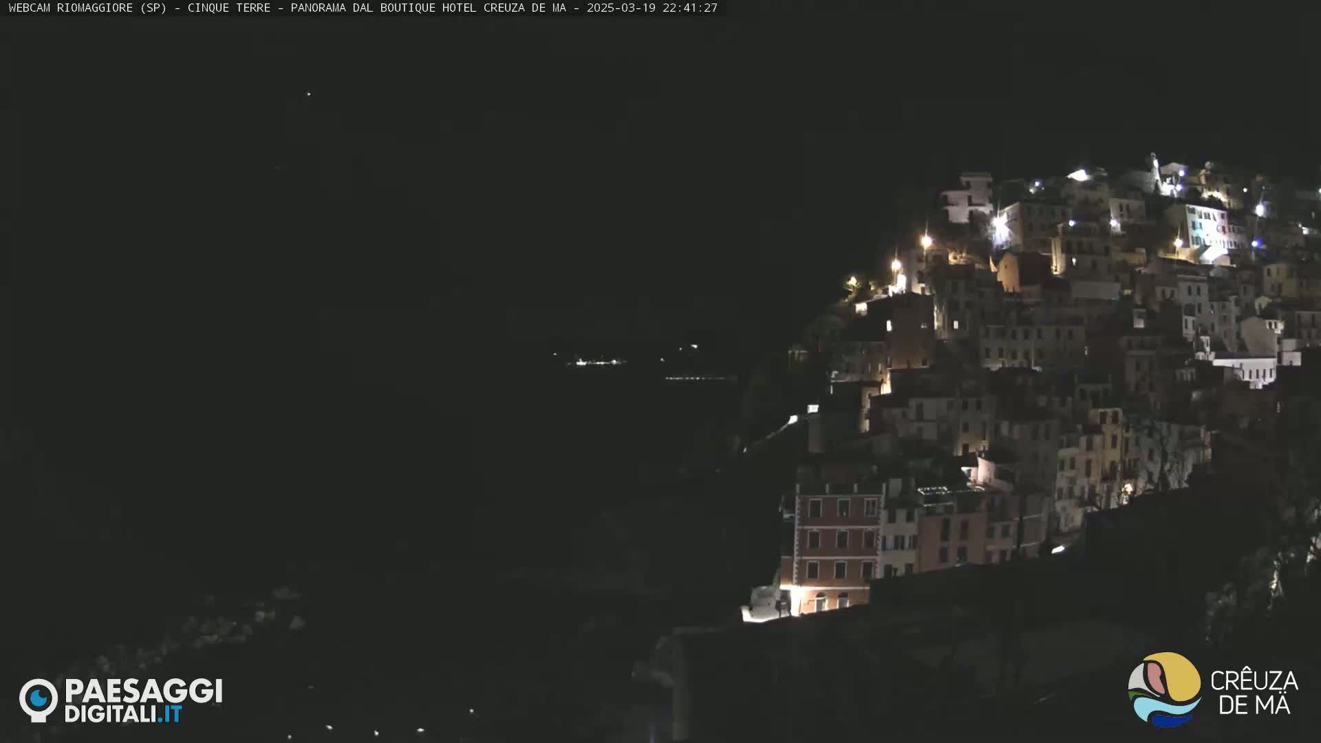 A nighttime view of a hillside town with many illuminated buildings, overlooking a dark body of water under a clear, dark sky.