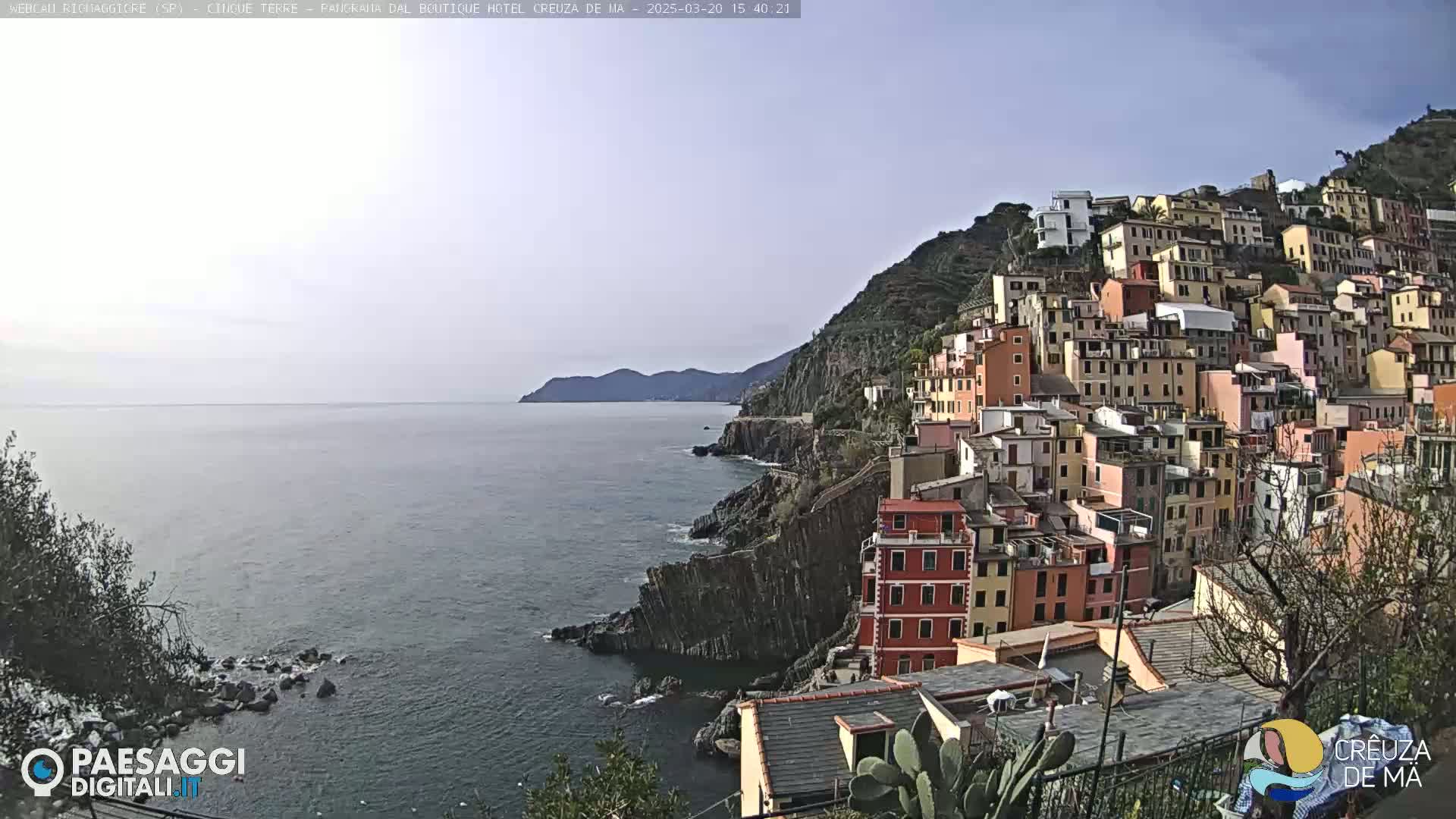 A coastal village of colorful buildings clings to a steep hillside overlooking a calm sea under a mostly clear sky.