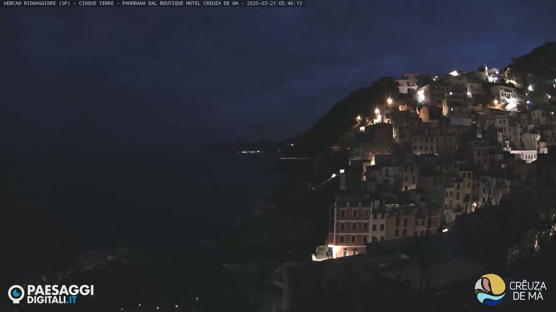 A nighttime view of a coastal town illuminated by lights, nestled on a hillside overlooking a dark body of water under a mostly clear, dark sky.