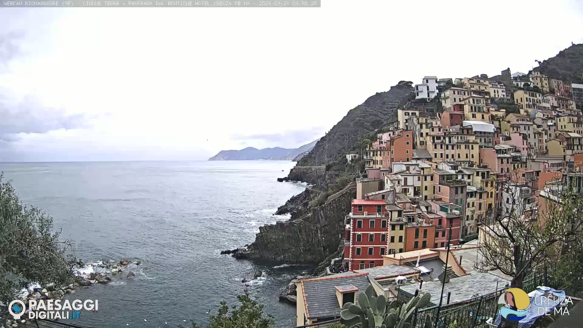 A coastal town of colorful buildings clings to a steep hillside overlooking a calm sea under a cloudy sky.