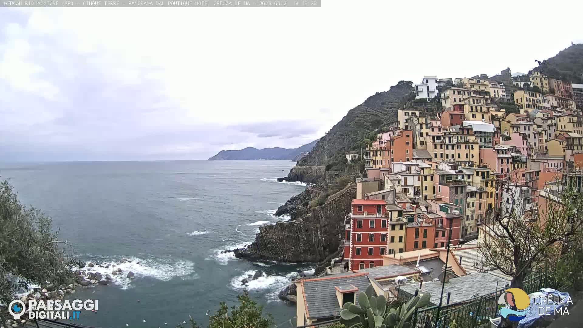 A coastal village of colorful buildings clings to a steep hillside overlooking a calm sea under an overcast sky.
