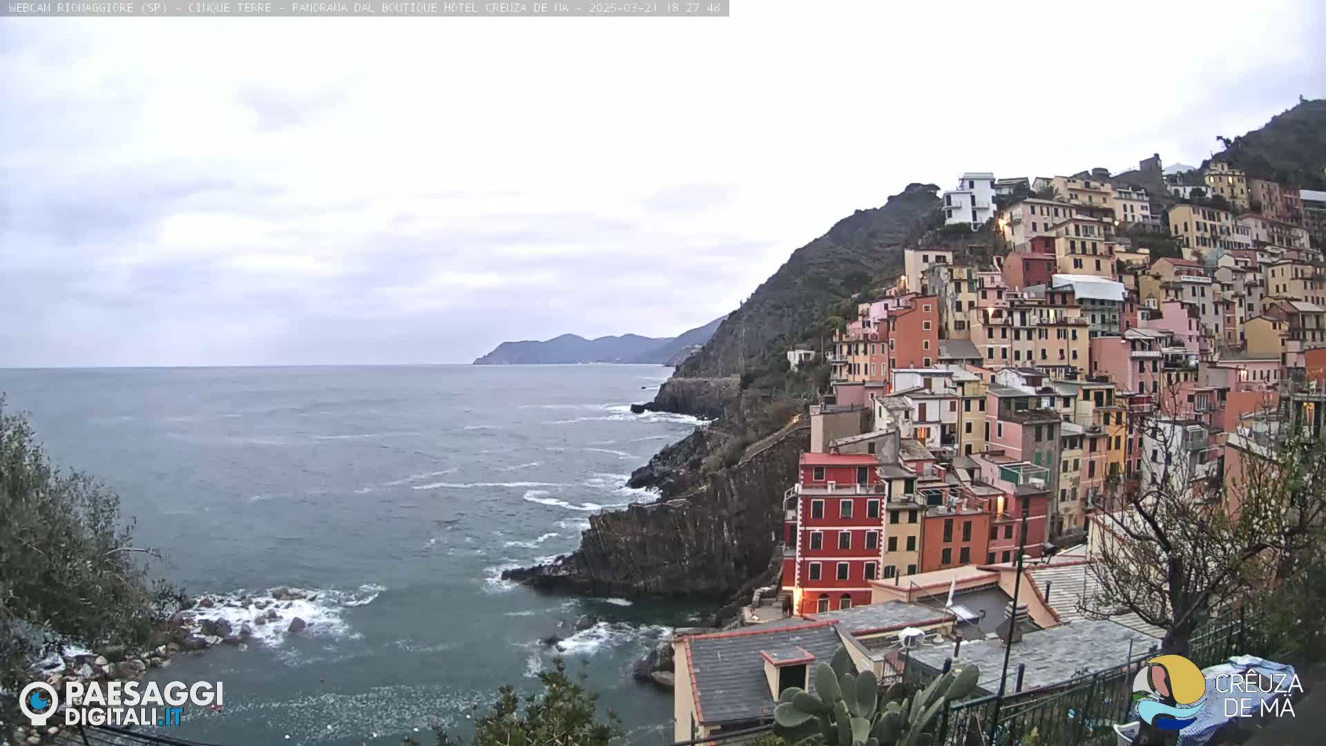 A coastal village of colorful buildings clings to a steep hillside overlooking a gray, somewhat choppy sea under an overcast sky.
