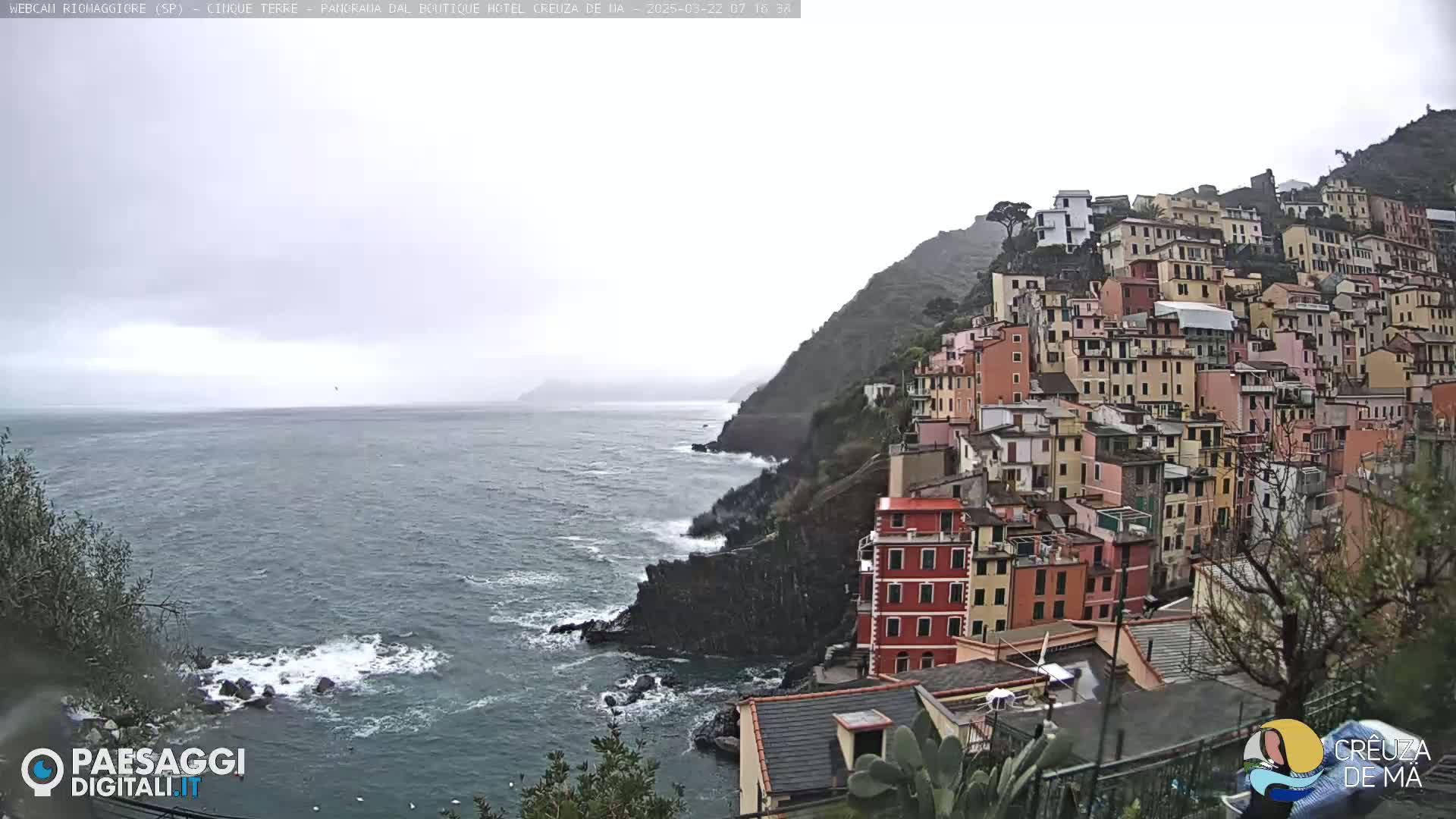 A coastal town of colorful buildings clings to a steep hillside overlooking a rough sea under an overcast sky.