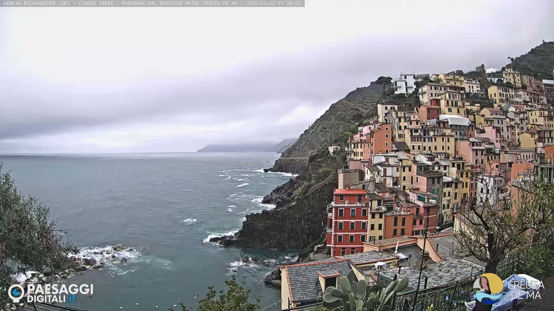 A coastal village of colorful buildings clings to a steep hillside overlooking a gray, overcast sea.