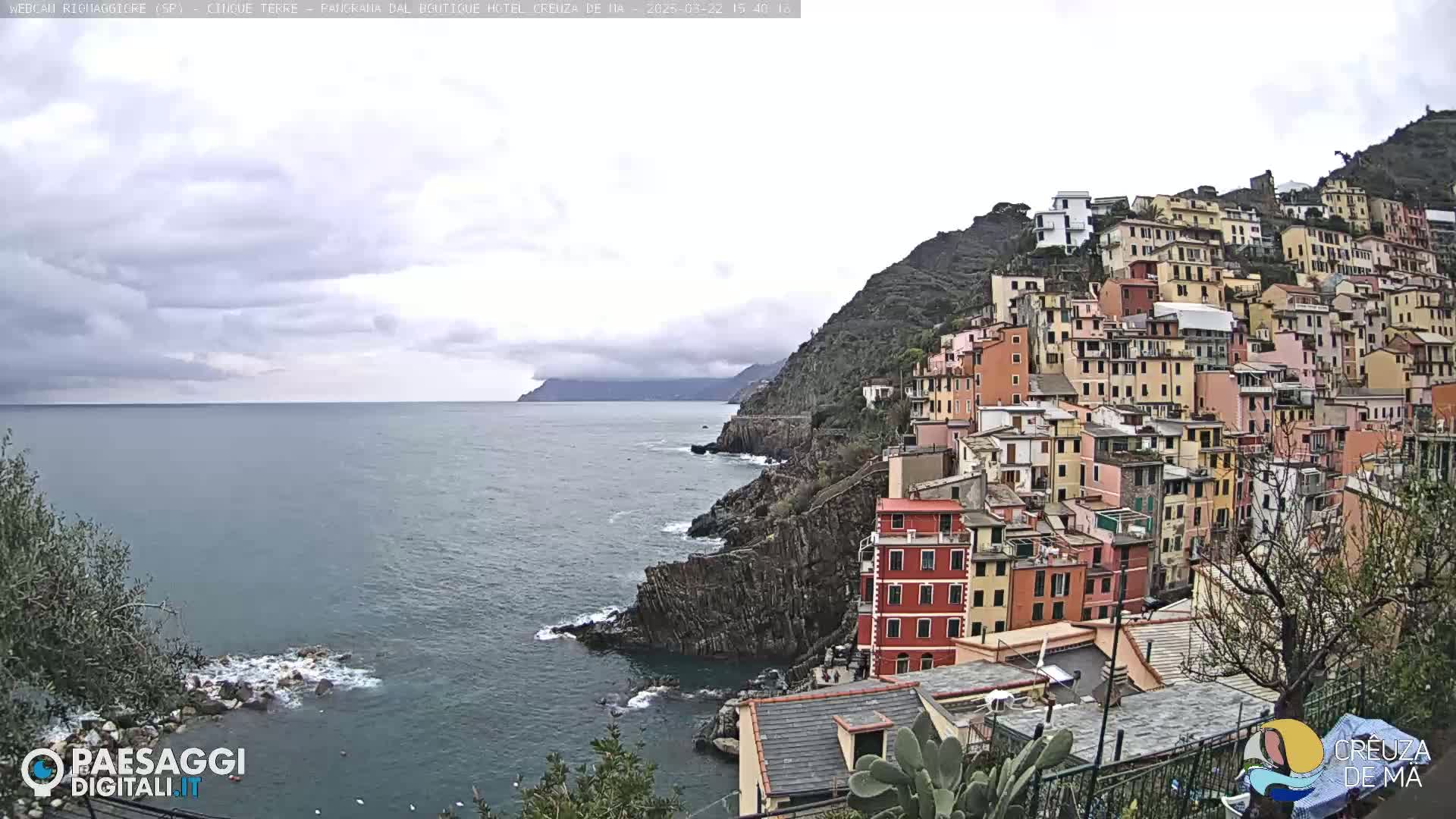 A coastal village of colorful buildings clings to a steep hillside overlooking a calm sea under a cloudy sky.