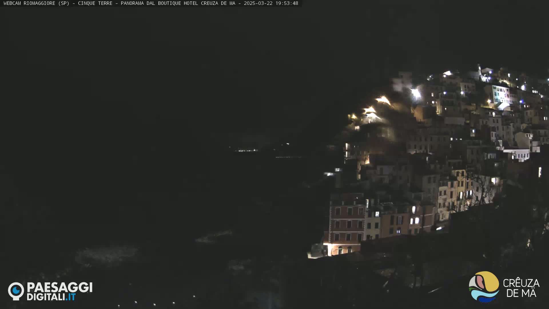 A nighttime view of a hillside town illuminated by lights, overlooking a dark body of water under a clear, dark sky.