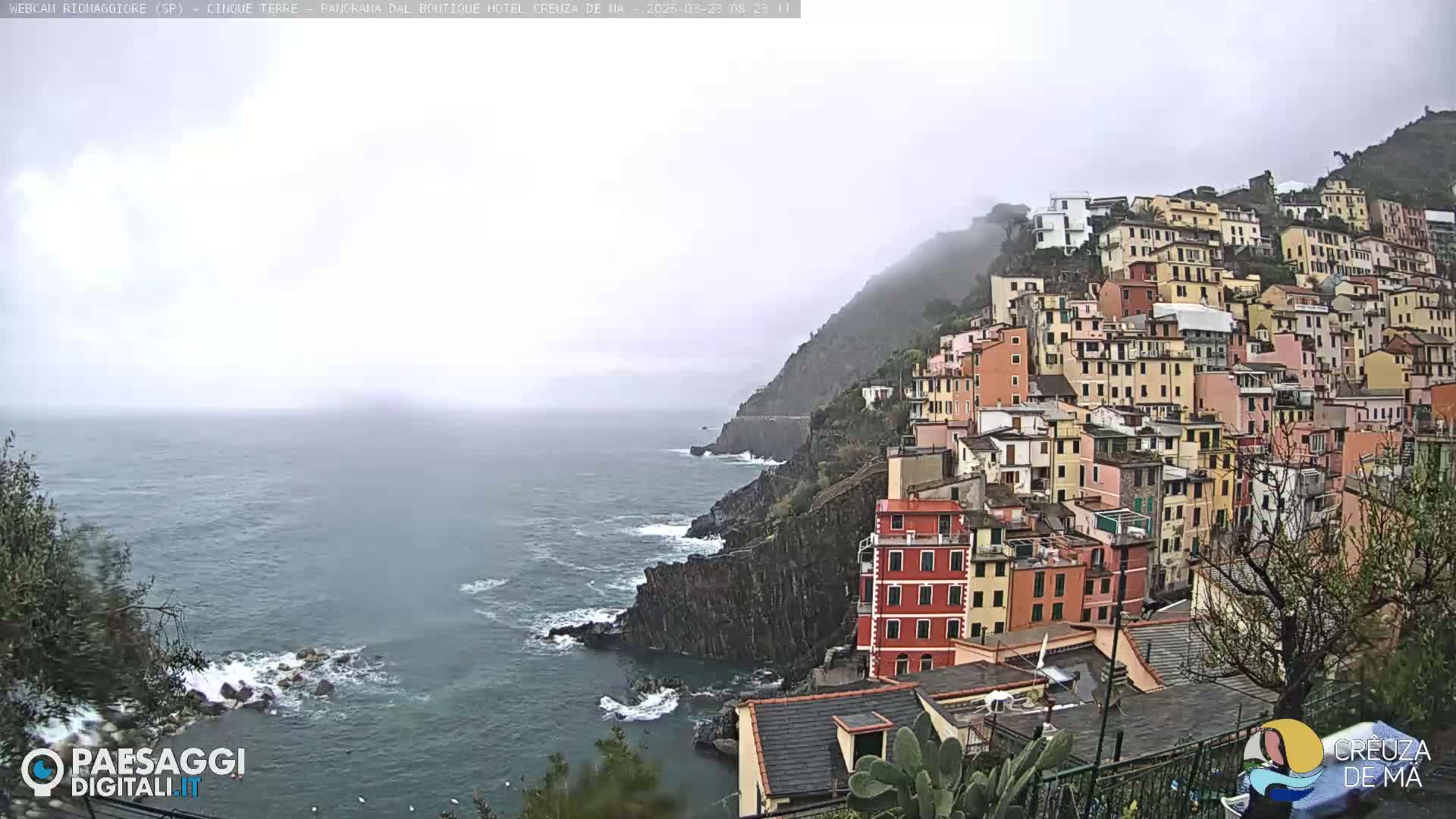 A coastal town of colorful buildings clings to a steep hillside overlooking a choppy sea under an overcast sky.