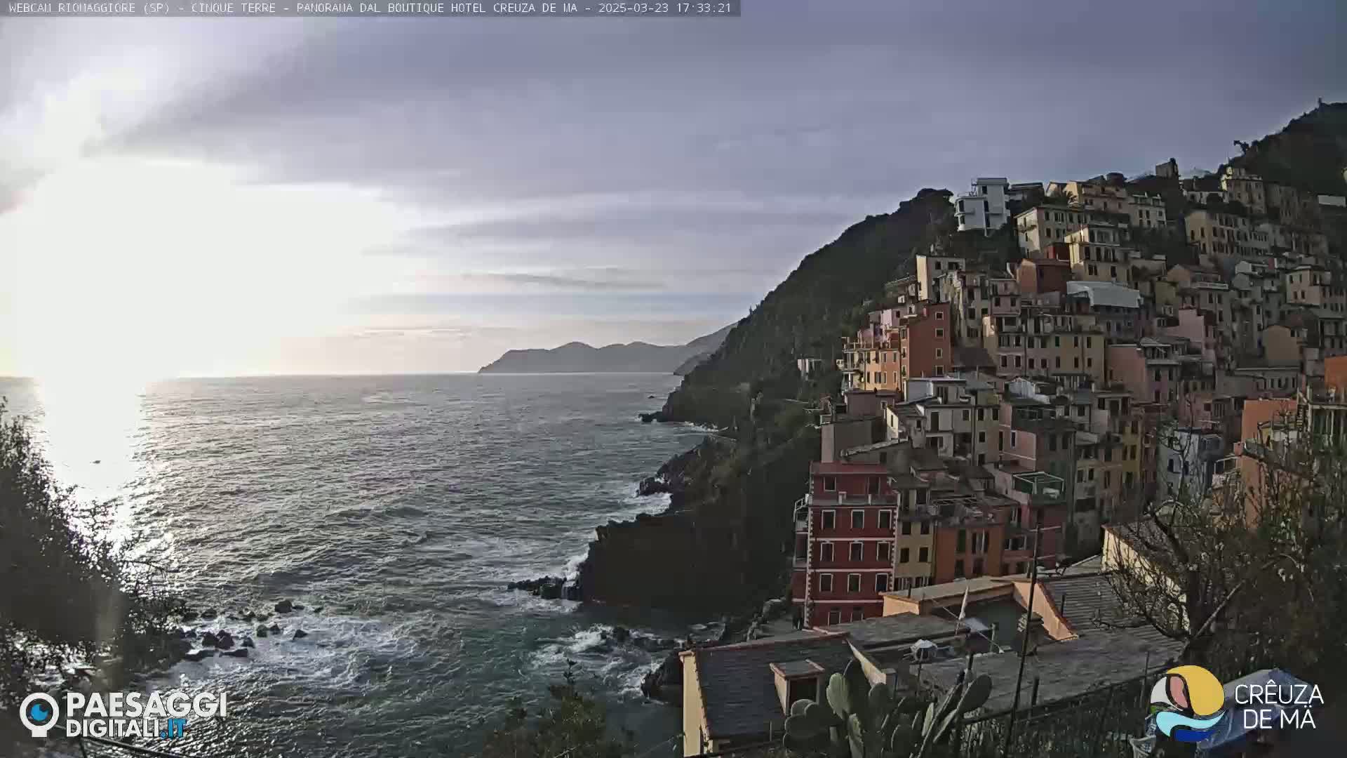 A coastal town with colorful buildings clings to a hillside overlooking a dark, choppy sea under a partly cloudy sky.