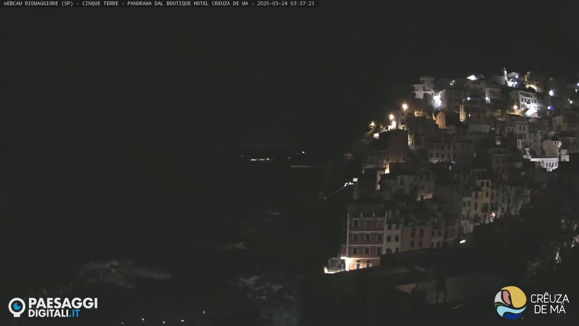 A nighttime view of a hillside town with illuminated buildings overlooking a dark body of water.