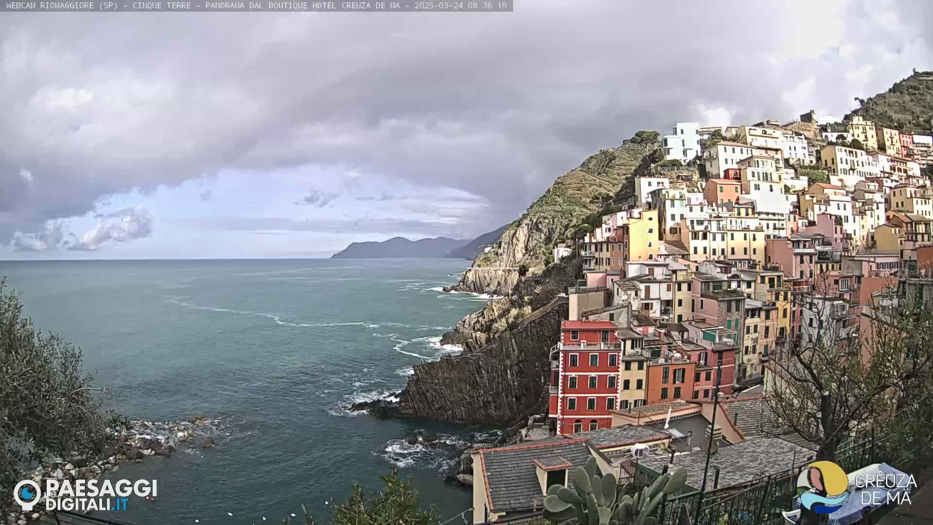 A colorful hillside village clings to a rocky coast overlooking a calm sea under a cloudy sky.