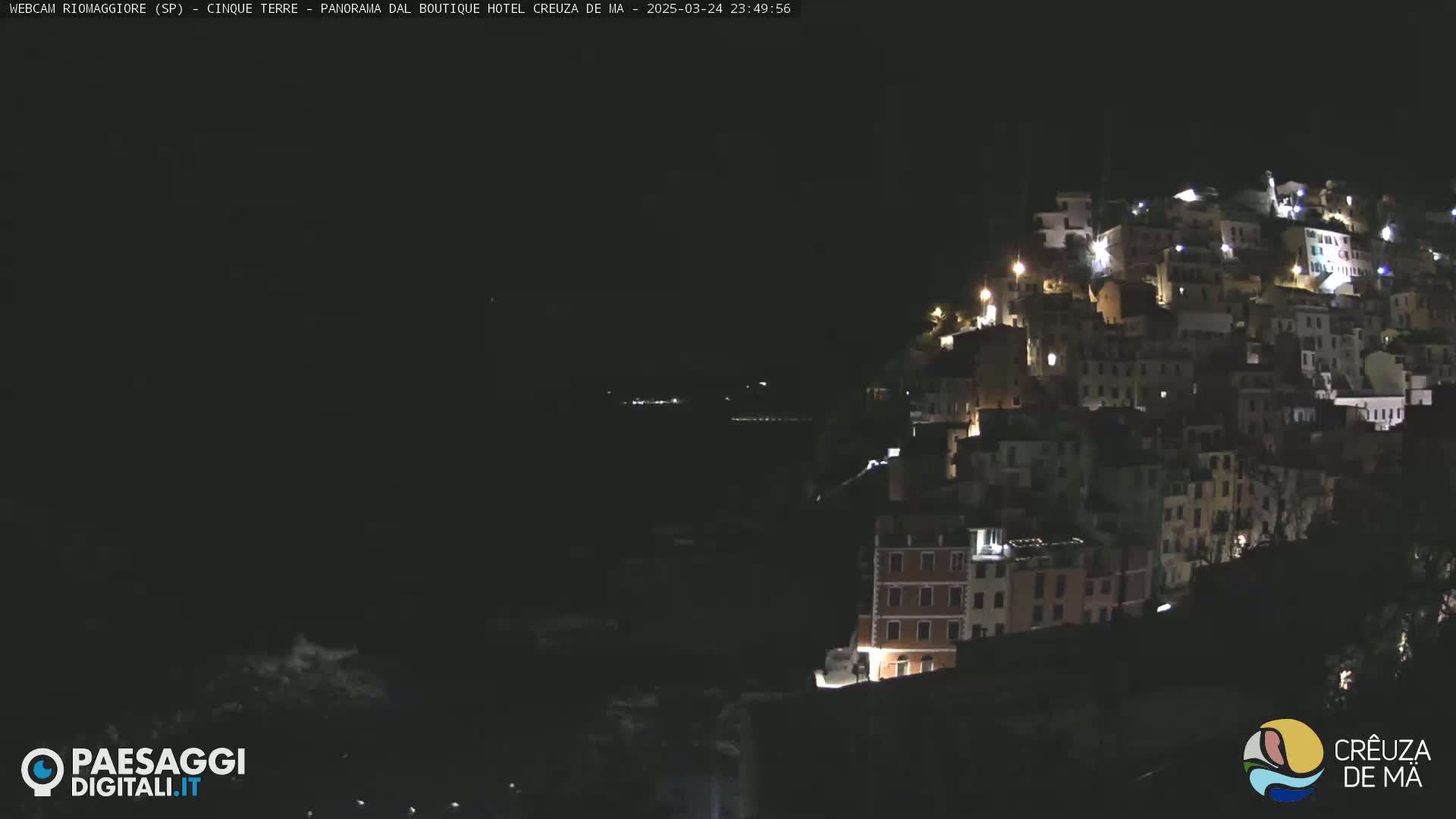 A nighttime view of a hillside town illuminated by lights, overlooking a dark body of water under a clear sky.