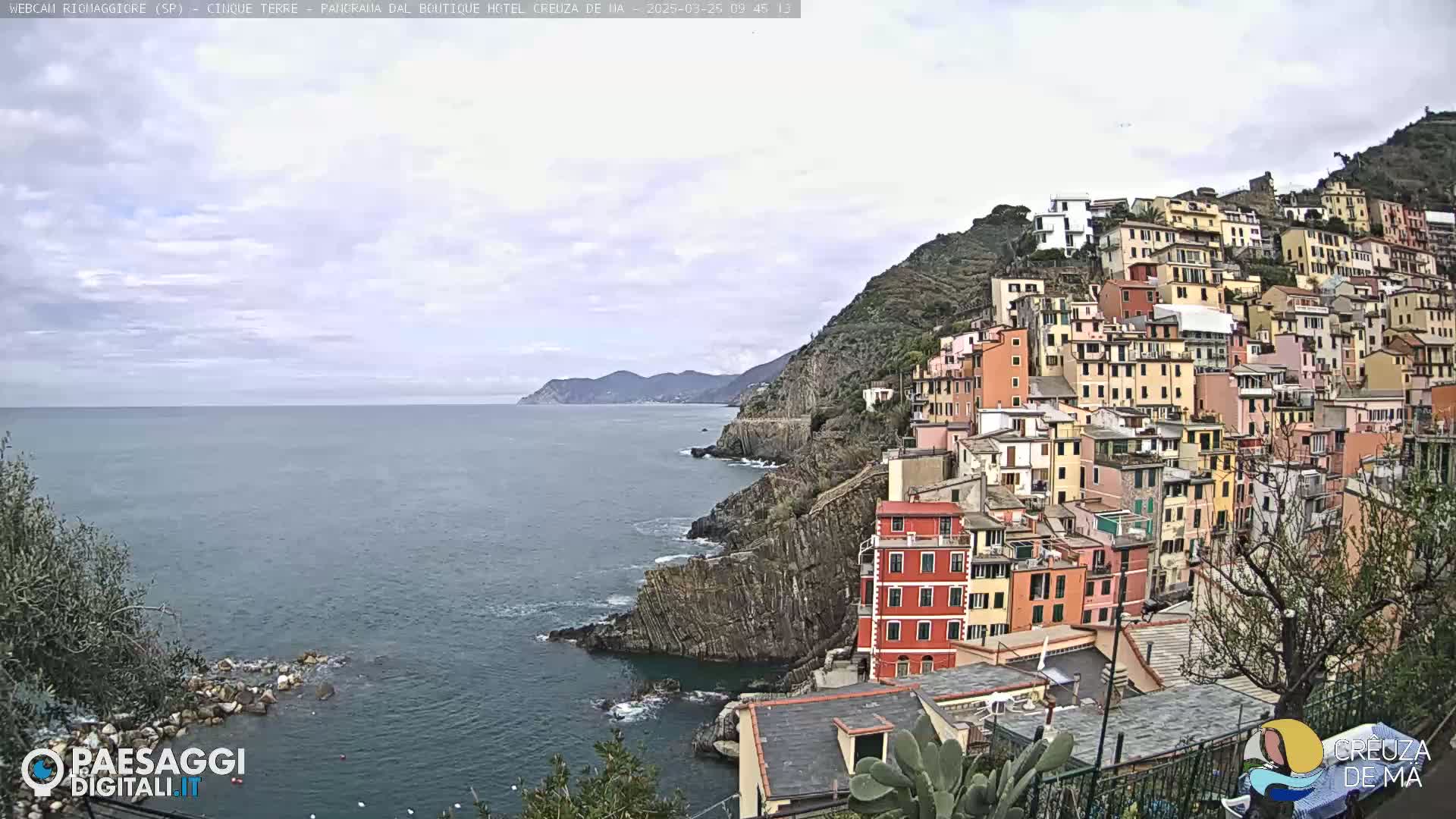 A coastal town of colorful buildings clinging to a steep cliffside overlooks a calm sea under a mostly cloudy sky.