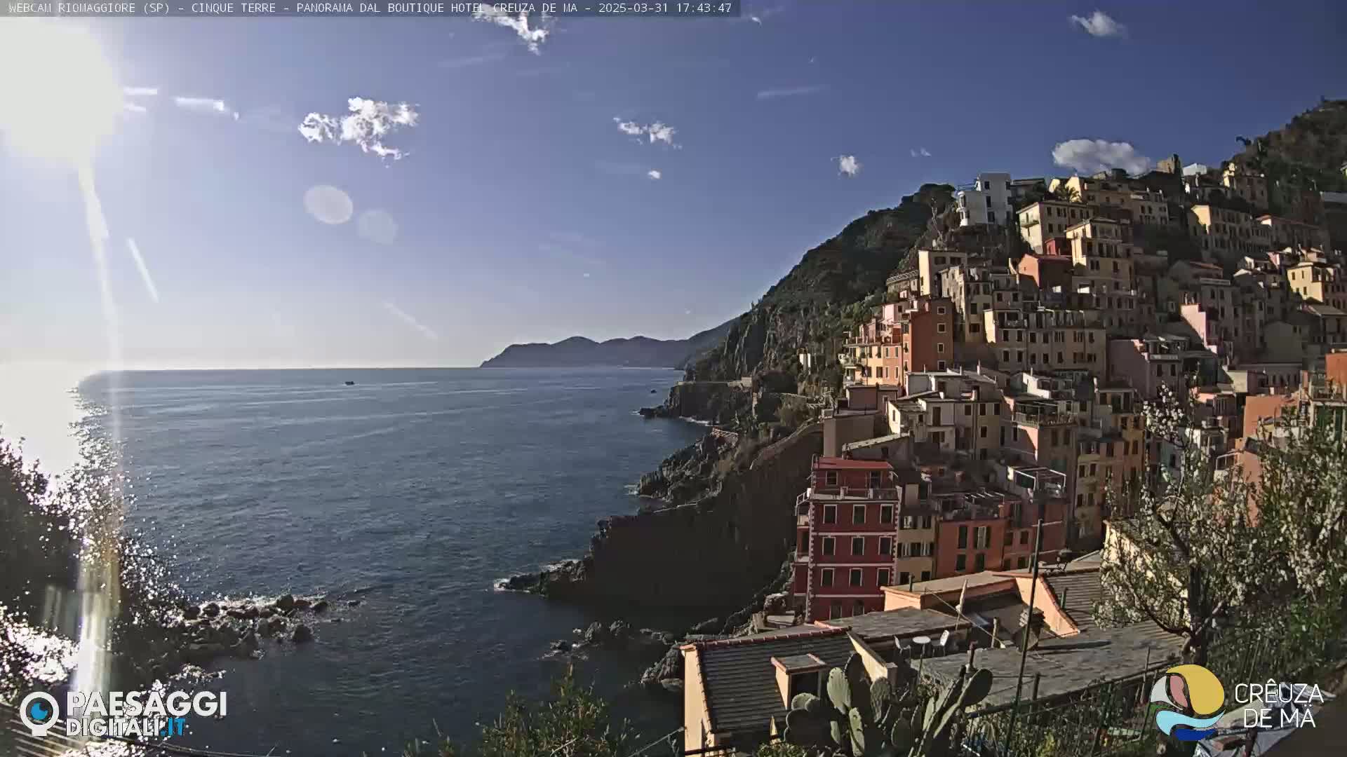 A colorful hillside village overlooks a calm, sunlit sea under a mostly clear sky.