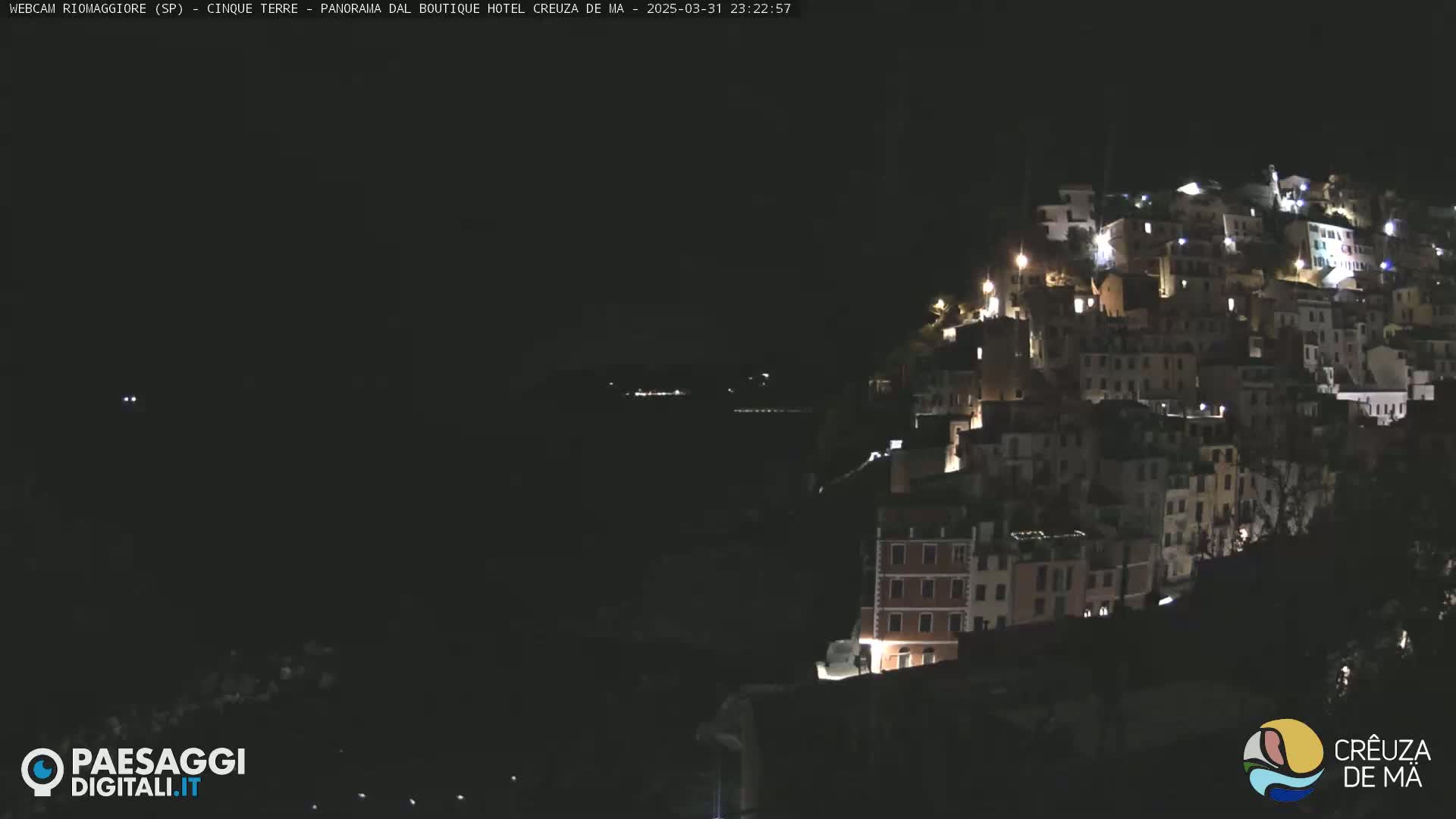 A nighttime view of a hillside village illuminated by lights, overlooking a dark body of water under a clear, dark sky.