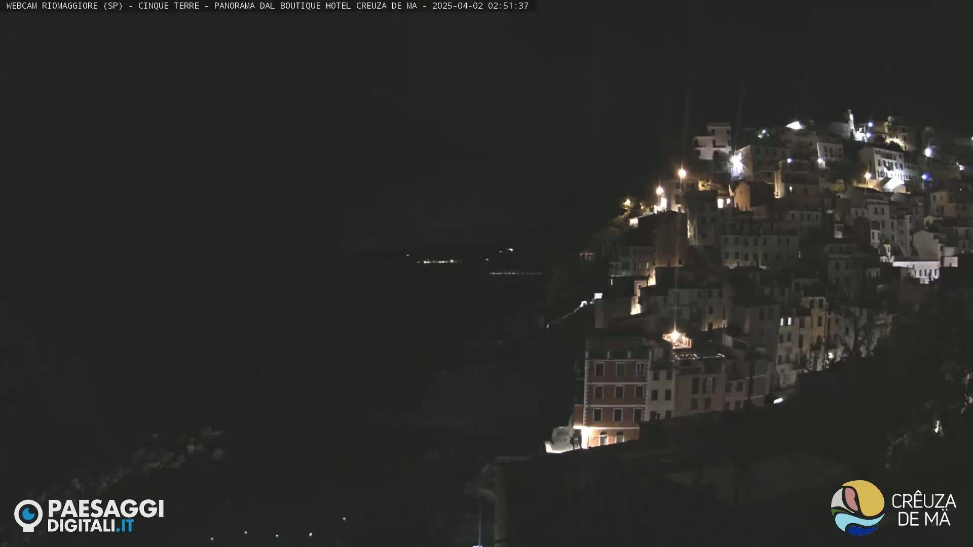 A nighttime view of a hillside town illuminated by lights, with a dark body of water visible in the foreground.