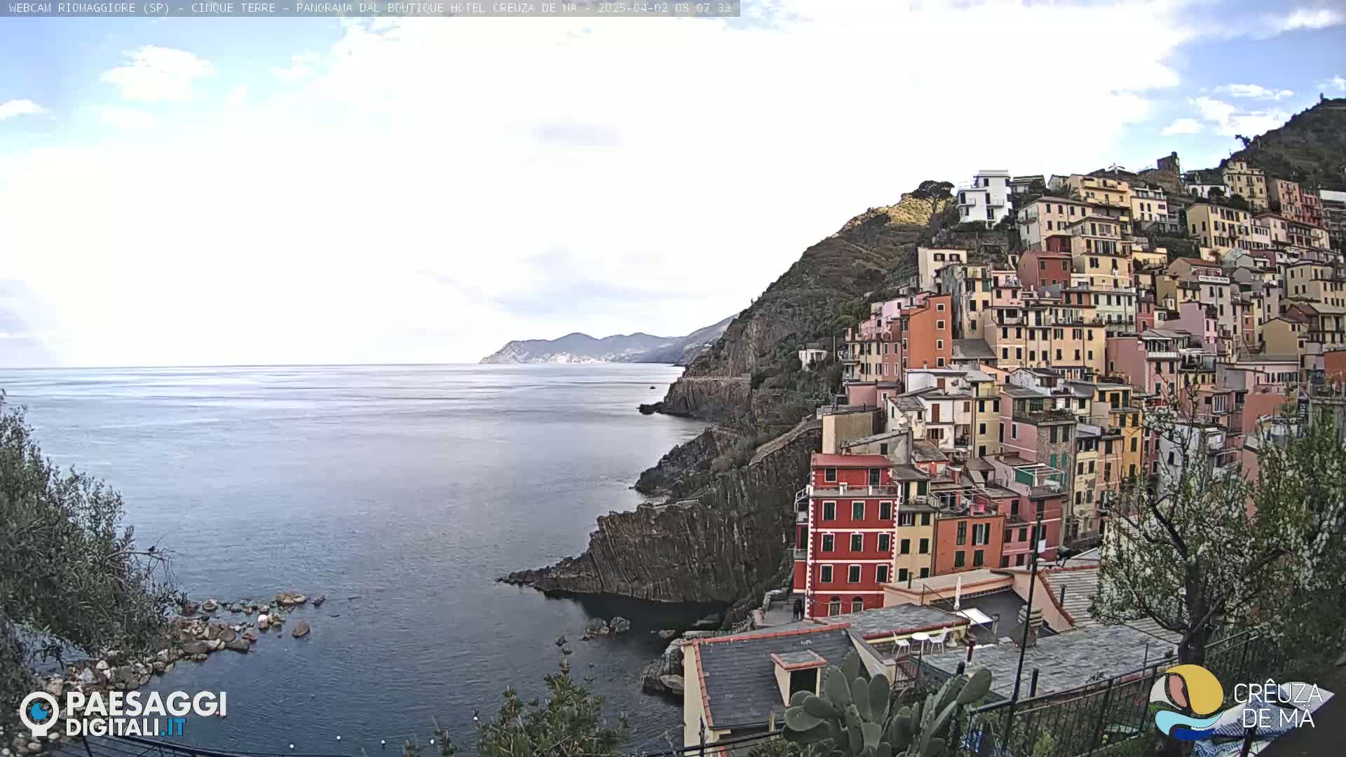 A coastal town of colorful buildings clings to a steep hillside overlooking a calm sea under a mostly cloudy sky.