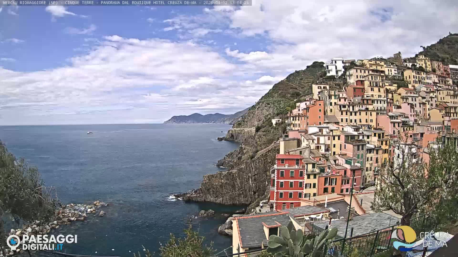 A colorful hillside village clings to a rocky coastline overlooking a calm sea under a partly cloudy sky.