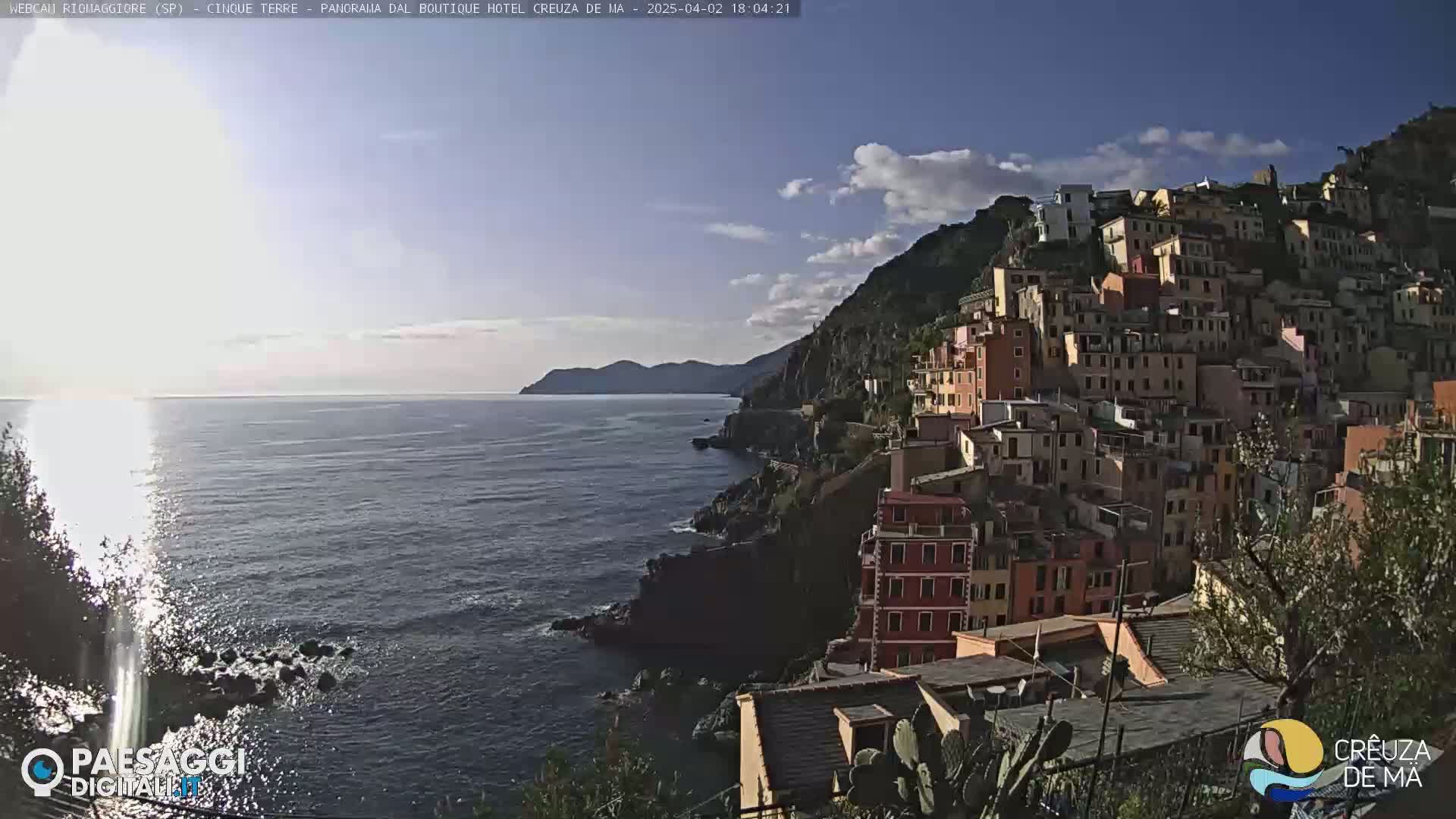 A coastal town with colorful buildings clinging to a steep hillside overlooks a calm sea under a partly cloudy sky.