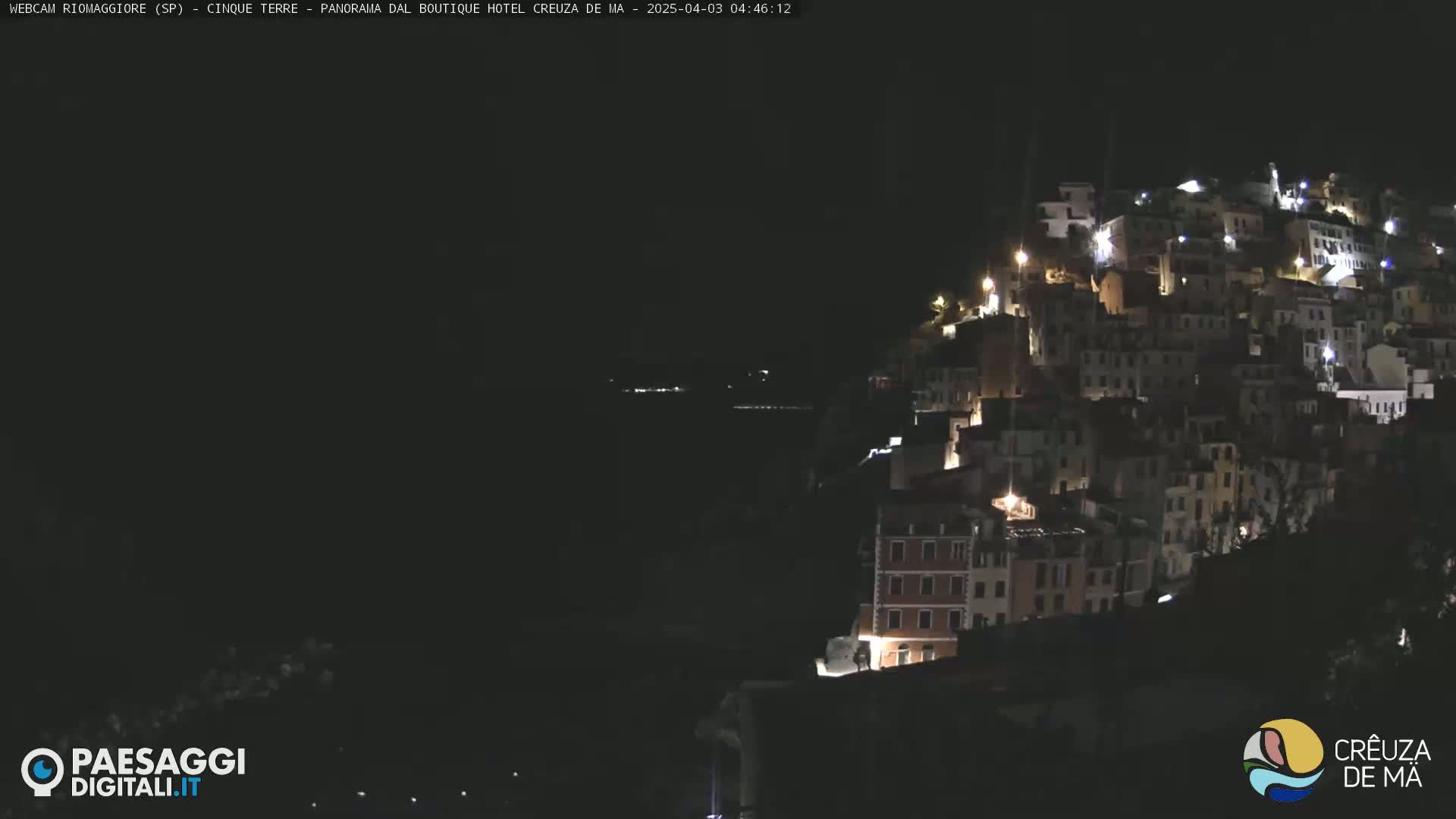 A nighttime view of a hillside town illuminated by lights, with a dark body of water visible in the distance.