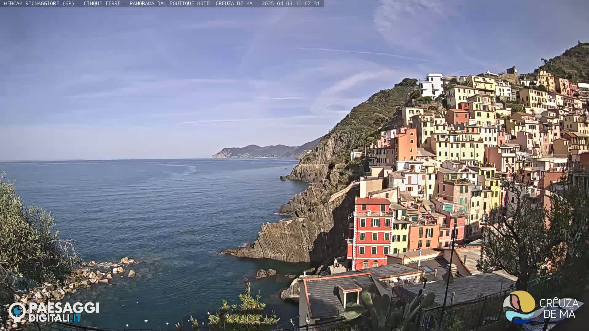 A colorful hillside village clings to a rocky coast overlooking a calm sea under a clear sky.
