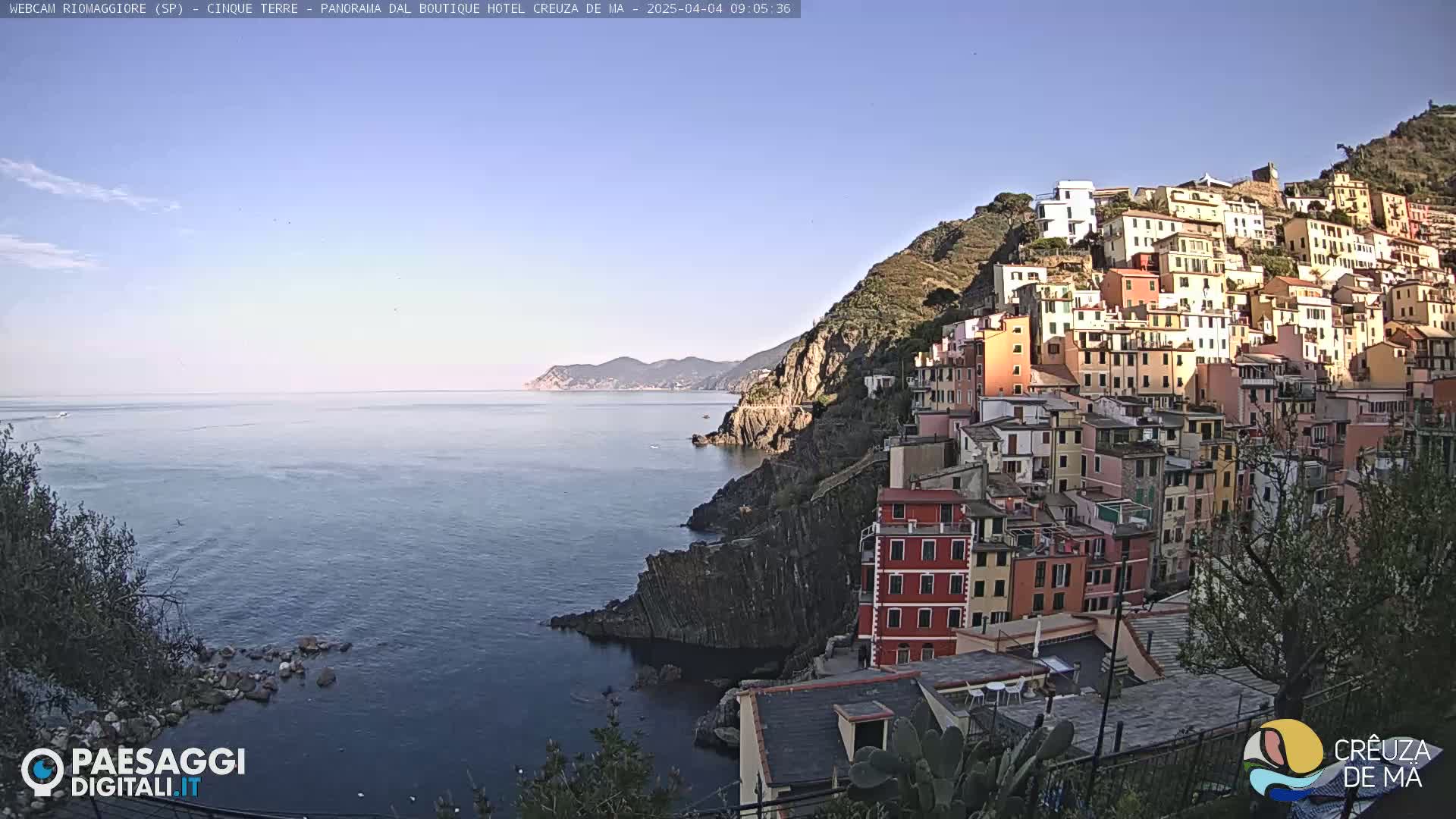 A colorful hillside village clings to a rocky coast overlooking a calm sea under a clear sky.